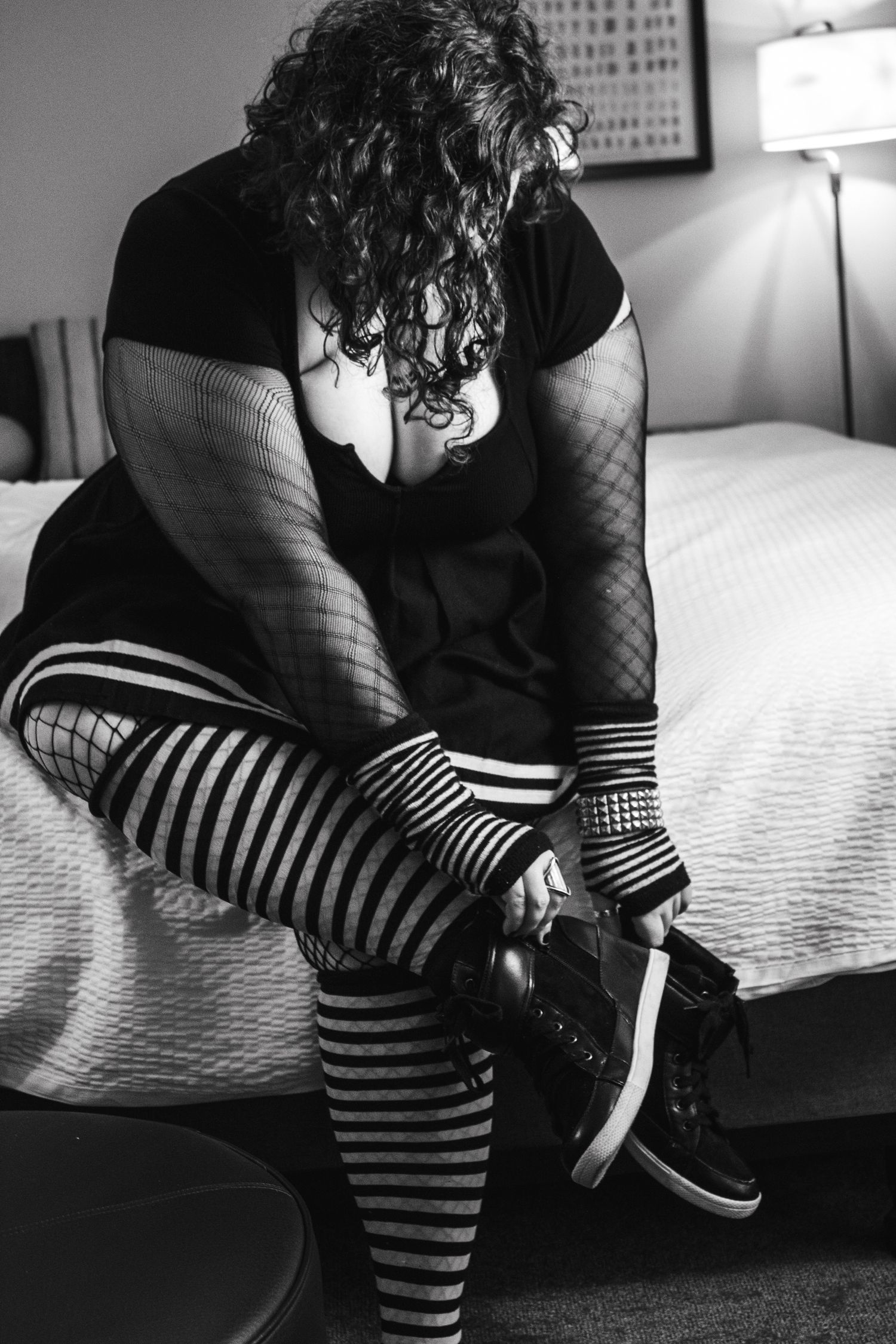 Person in black outfit and striped socks sits on bed putting on shoes.
