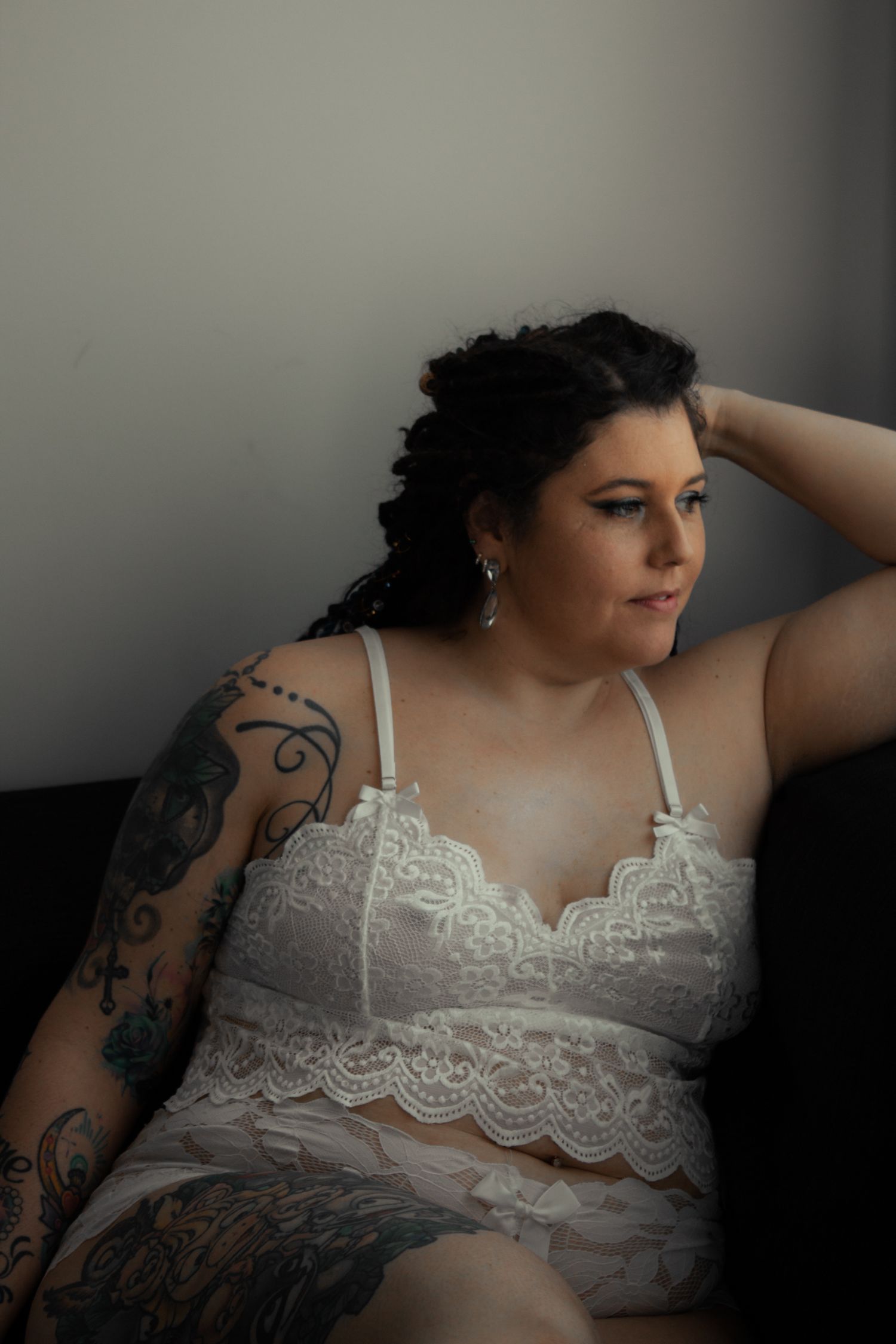 Person in white lace lingerie with tattoos poses against grey wall.