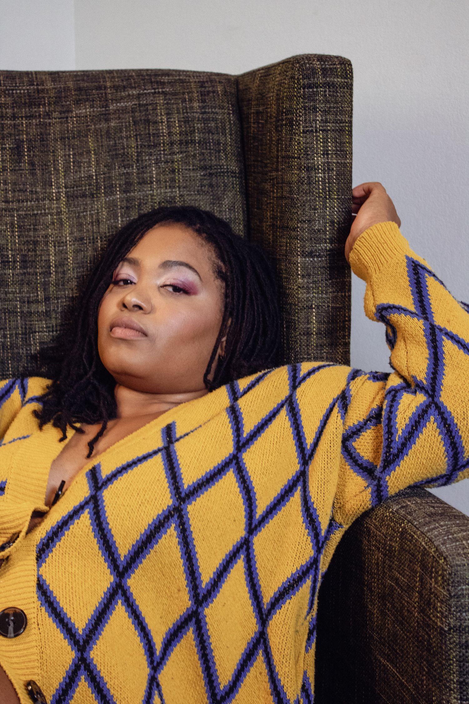 Model wearing bright yellow and navy diamond pattern sweater poses dramatically on couch.