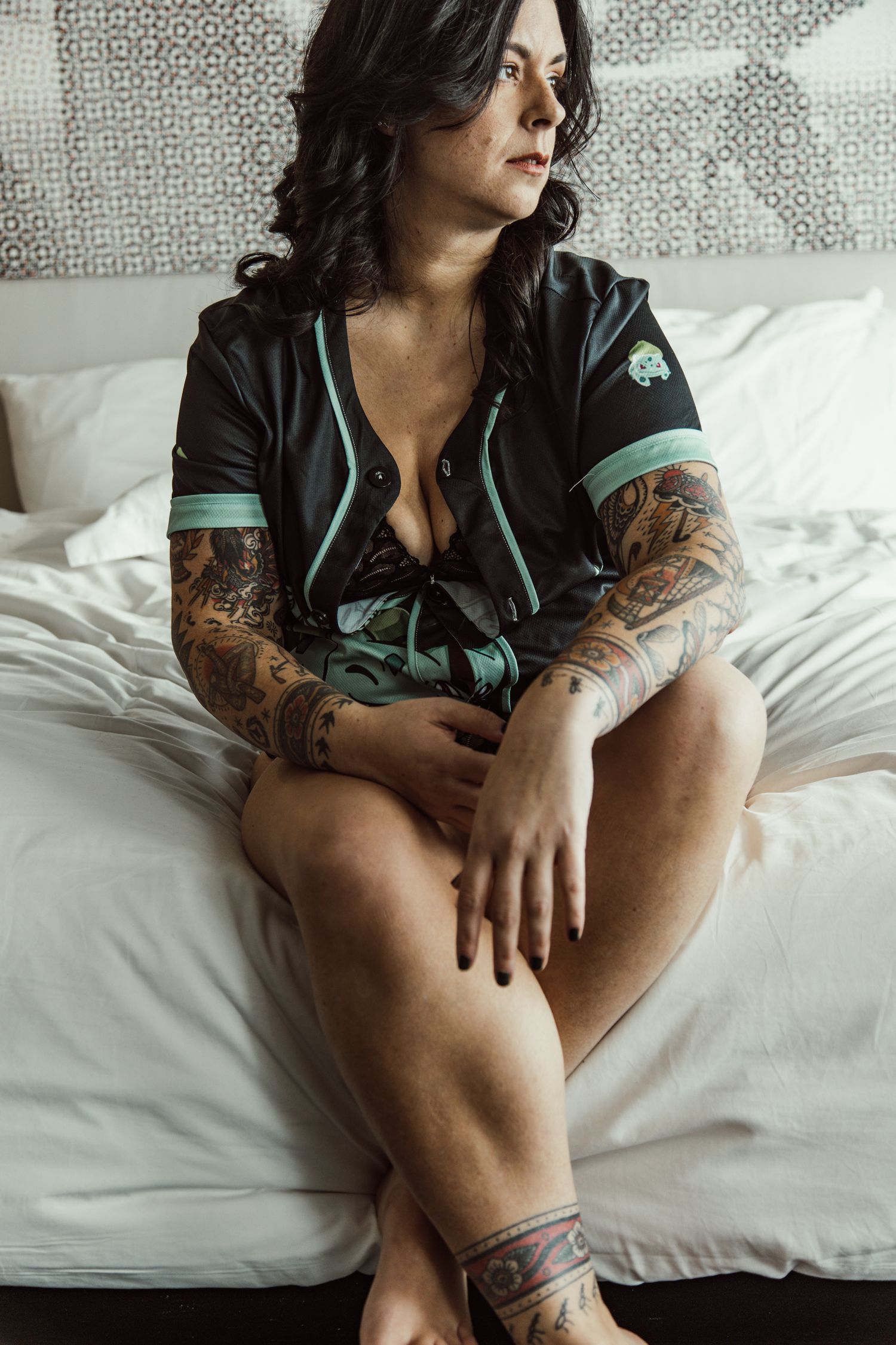 Person with tattoos poses casually on white bedding wearing black and teal.