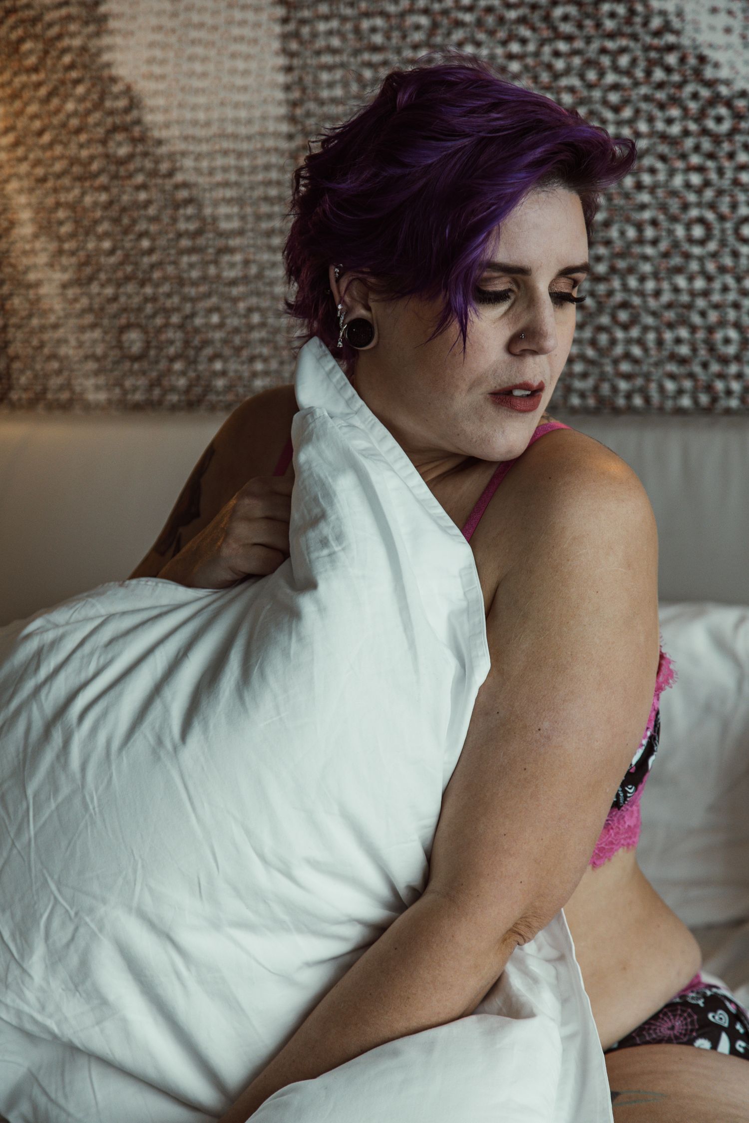 Person with purple hair lying on white bedding in intimate boudoir photoshoot.