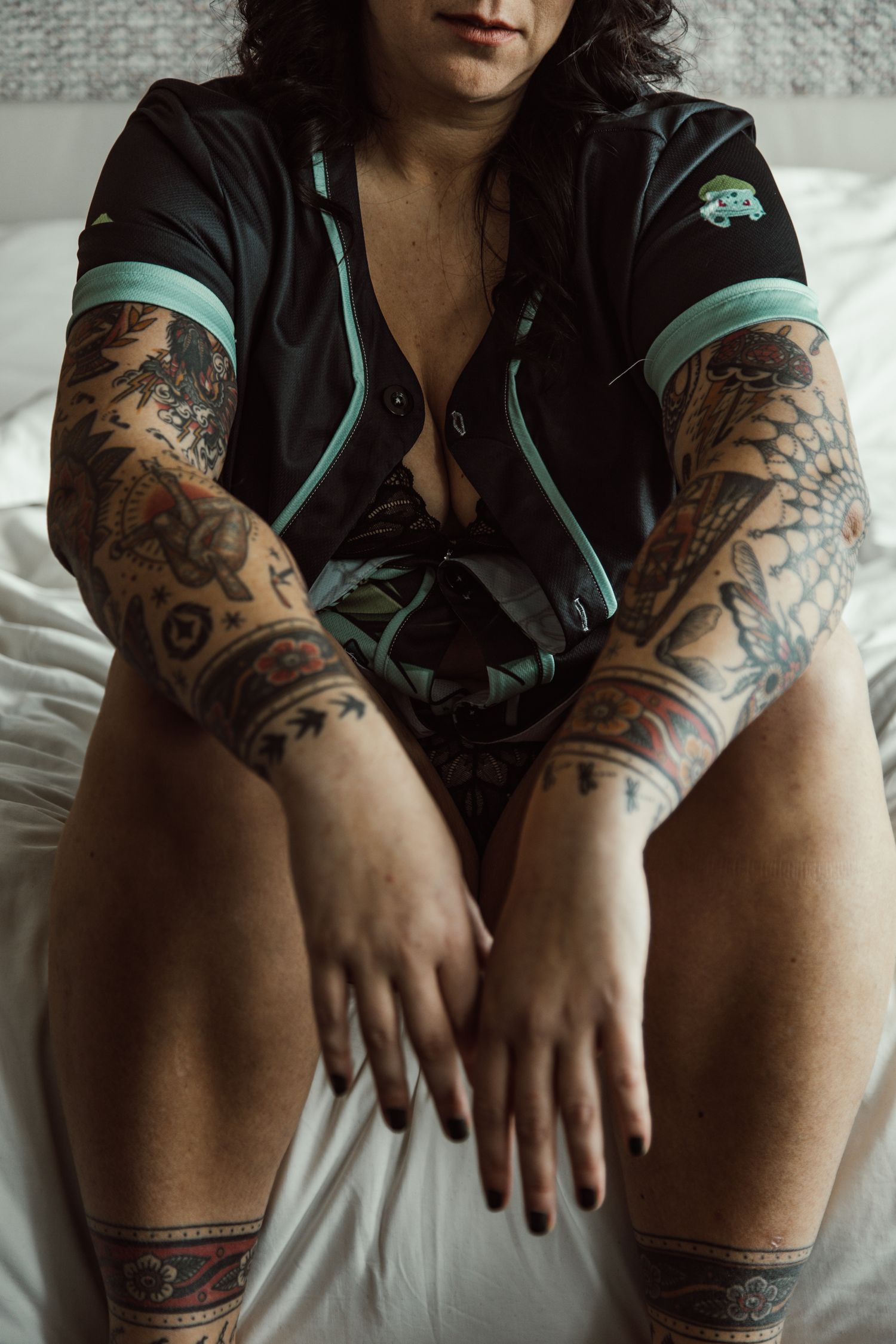 Person with tattoo sleeves sits on white bed wearing black and teal clothing.