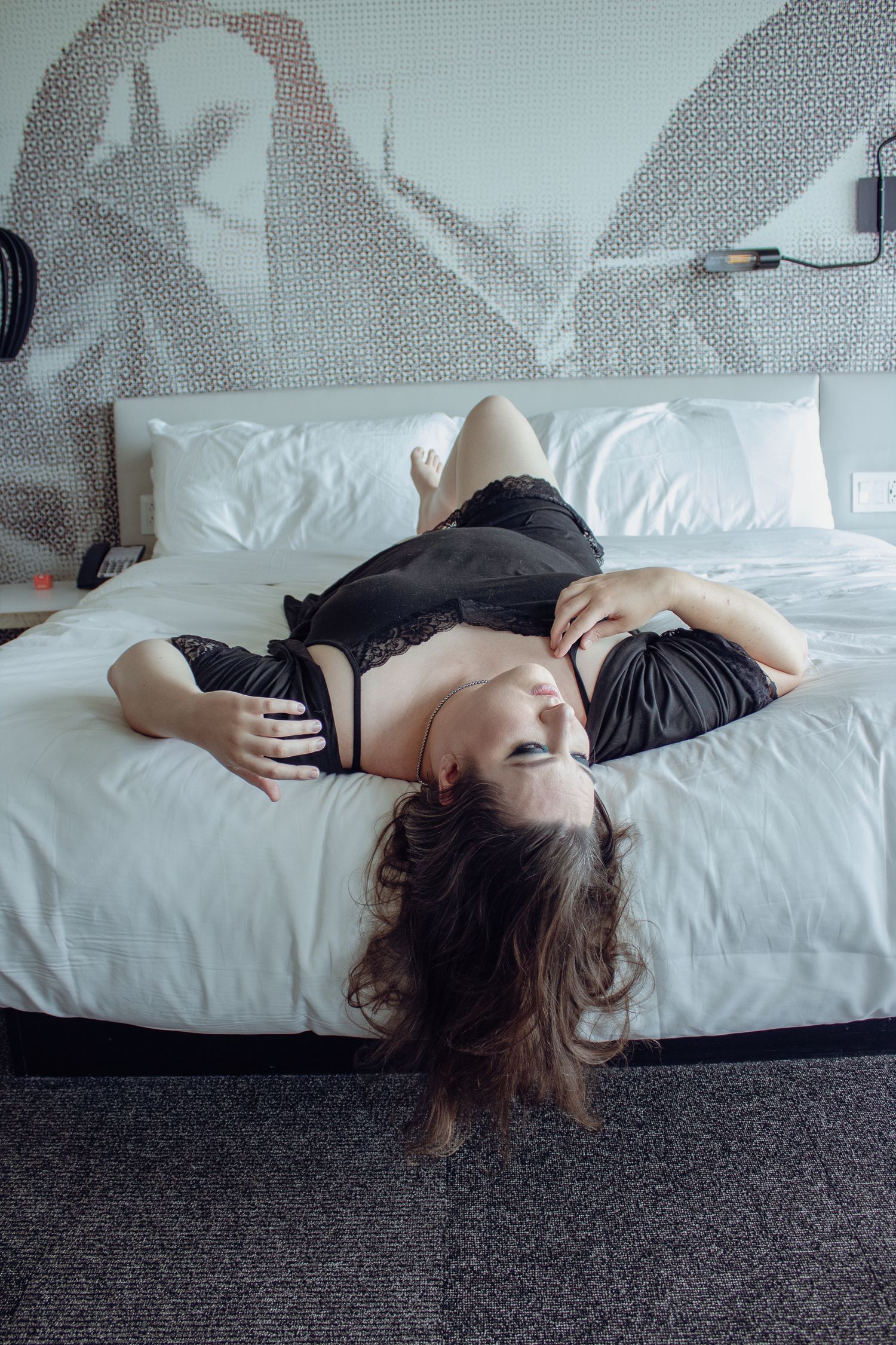 Person in black lingerie lying on white bed with artistic wall pattern behind.