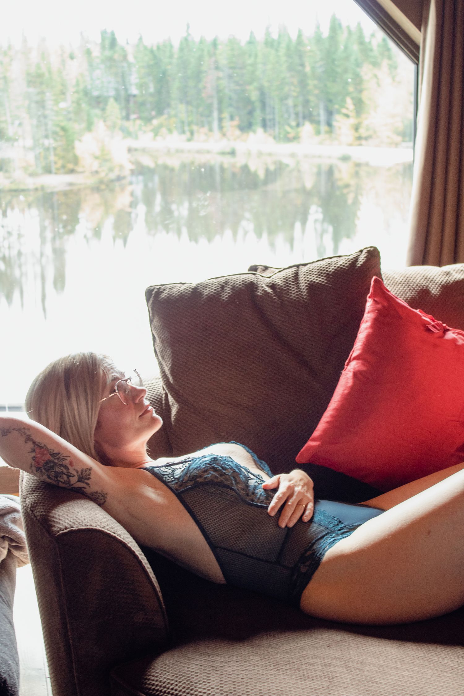 Person relaxing on couch by window overlooking serene lake and forest landscape.