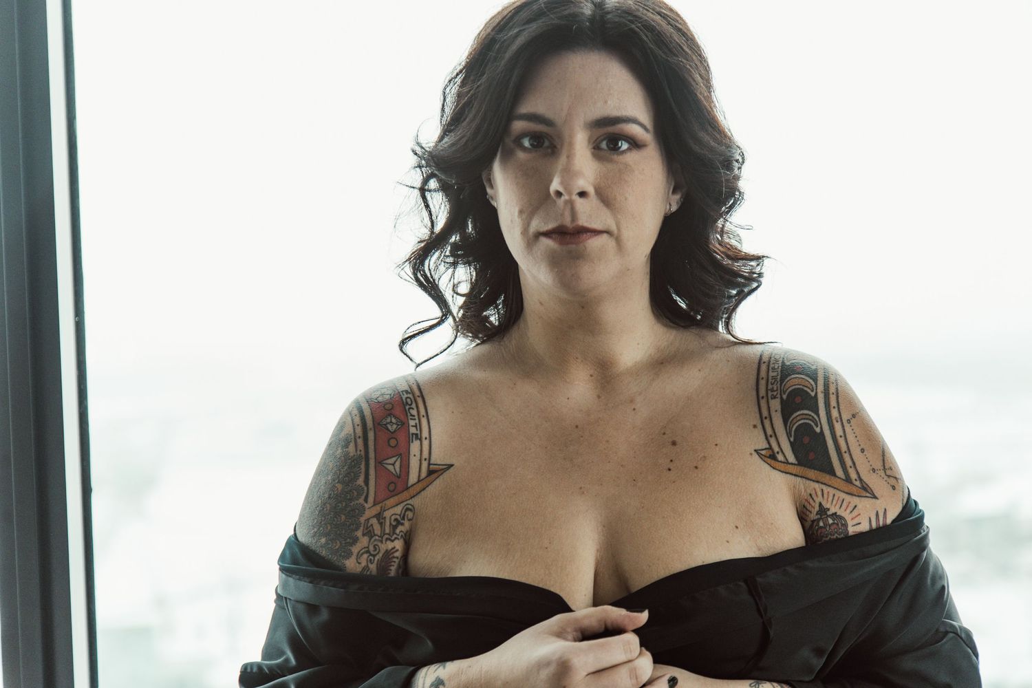 Moody portrait against bright window showing shoulders with visible tattoo art.