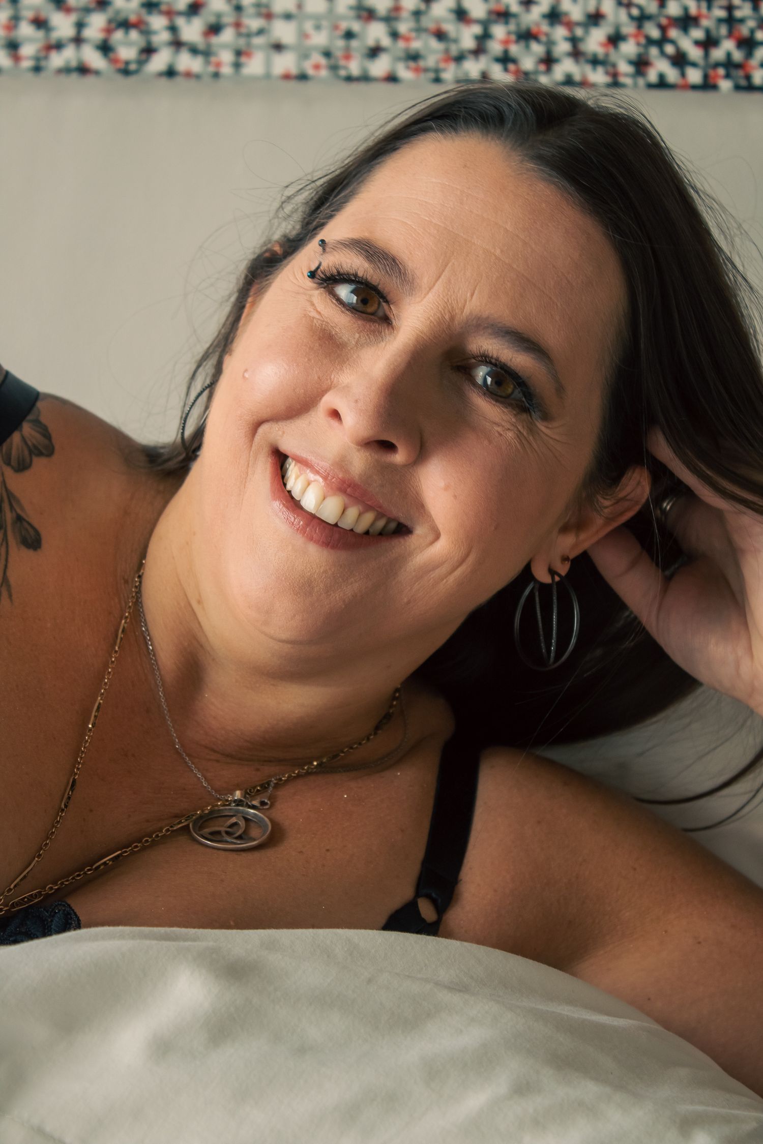 Person with dark hair and tattoos smiling while reclining on bed.