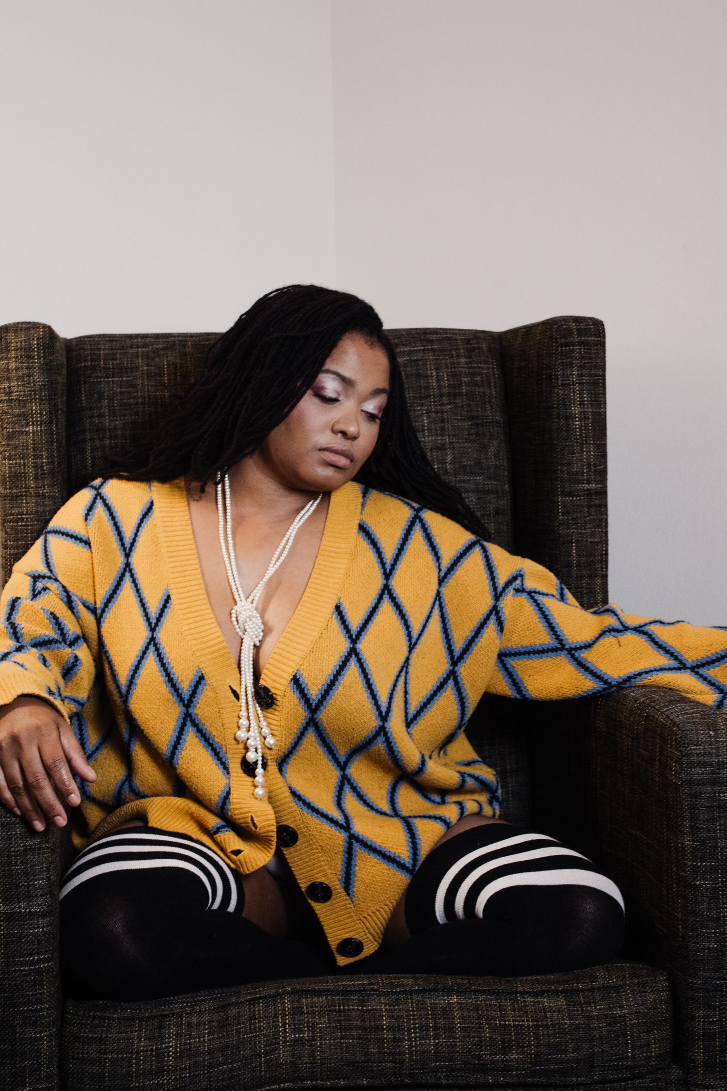 Model wearing yellow and blue patterned sweater lounges on dark sofa.