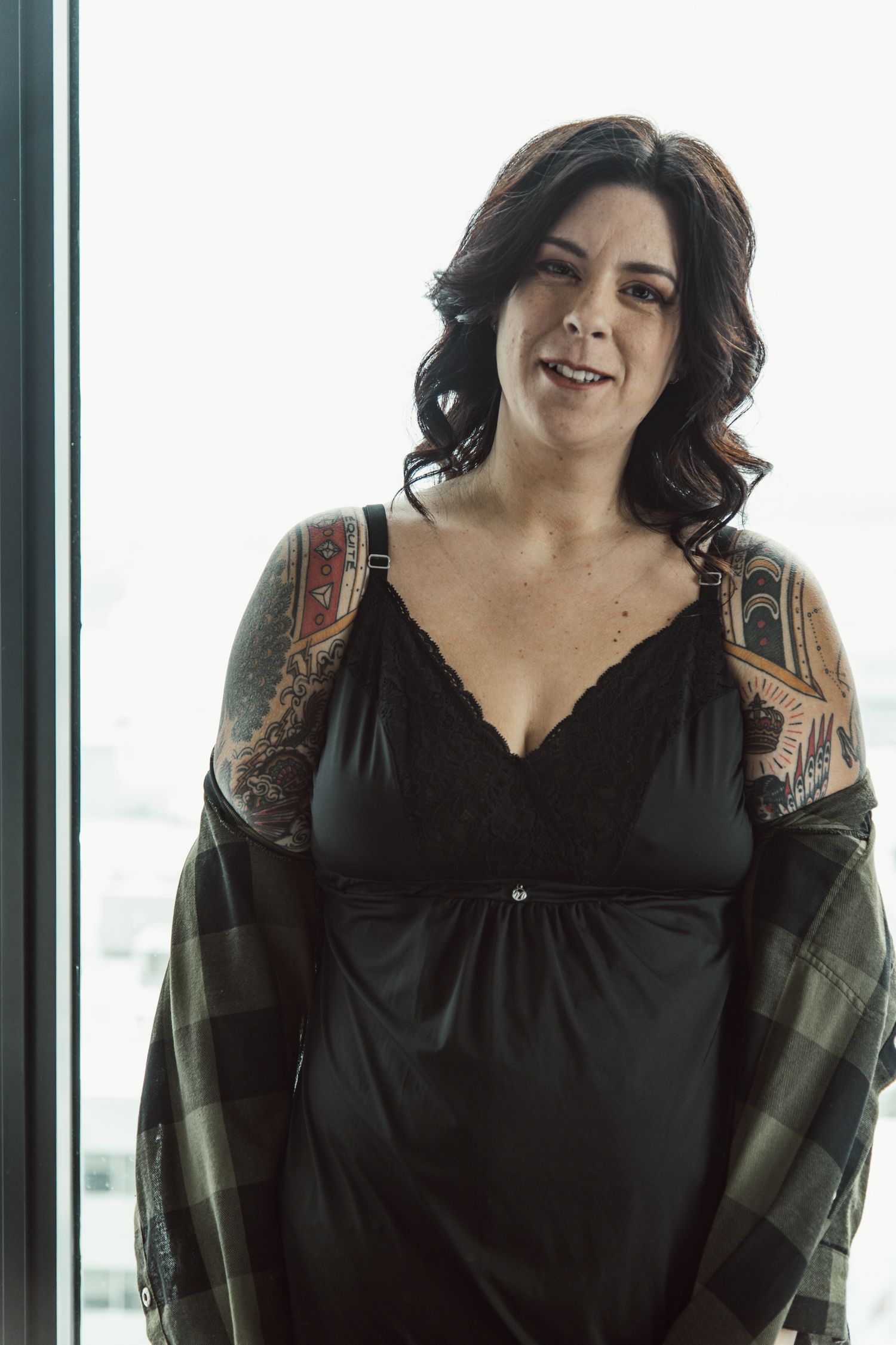 Person wearing black top and plaid shirt poses by window in casual photoshoot.