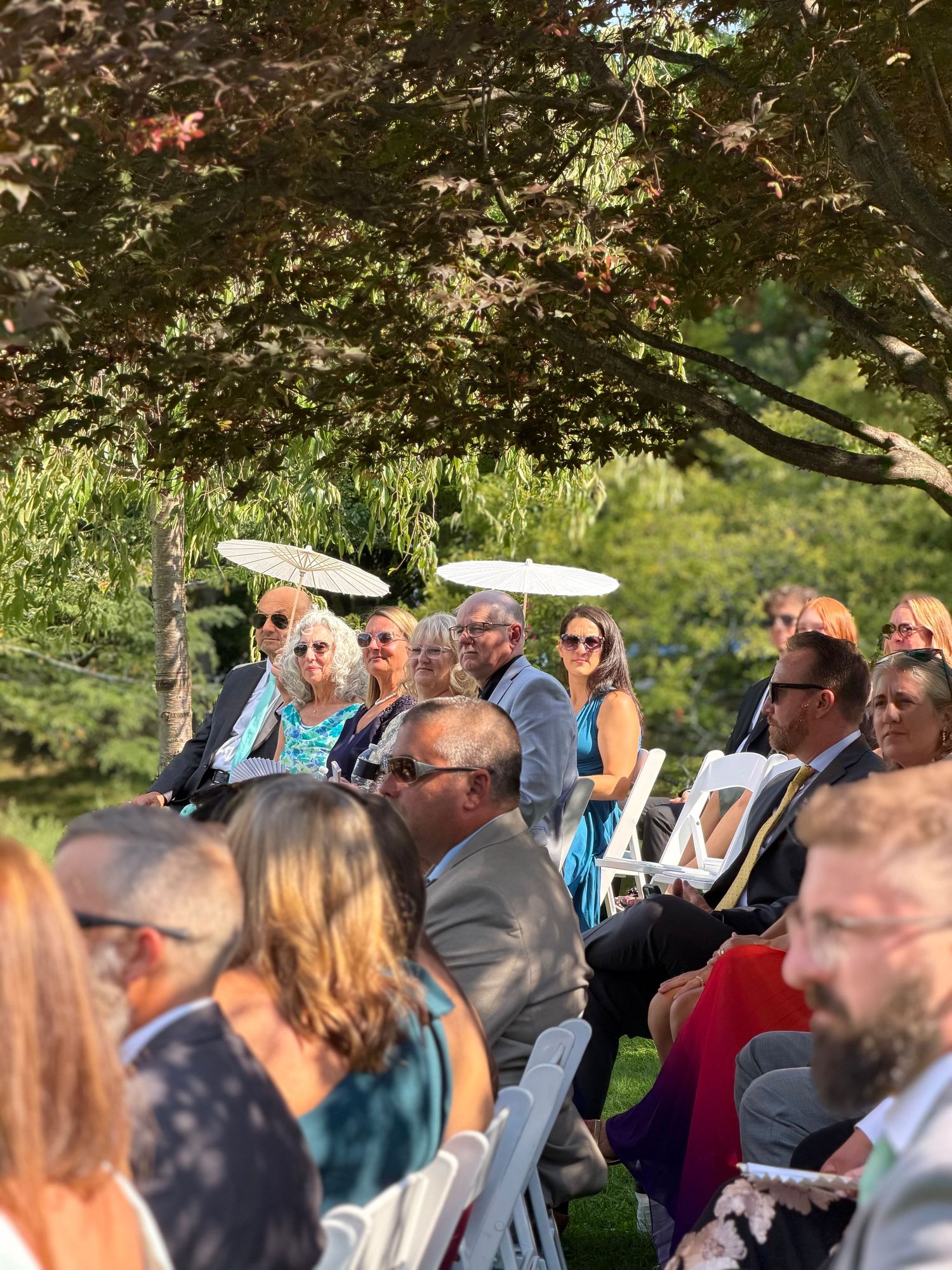 Guests seated in white chairs at an outdoor wedding ceremony under lush green trees on a sunny day.