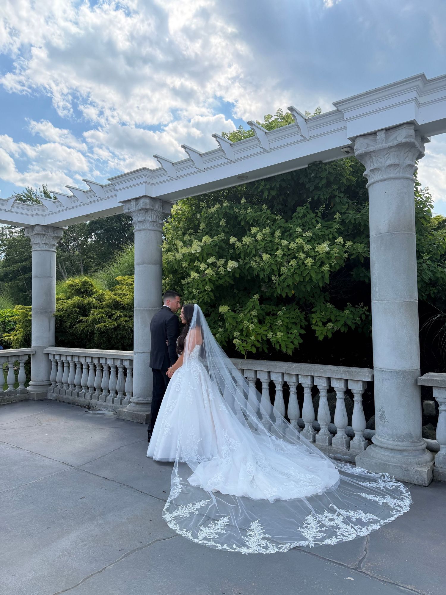 A bride and groom share a romantic moment under an elegant white pergola with marble columns, her lace cathedral veil trailing beautifully.