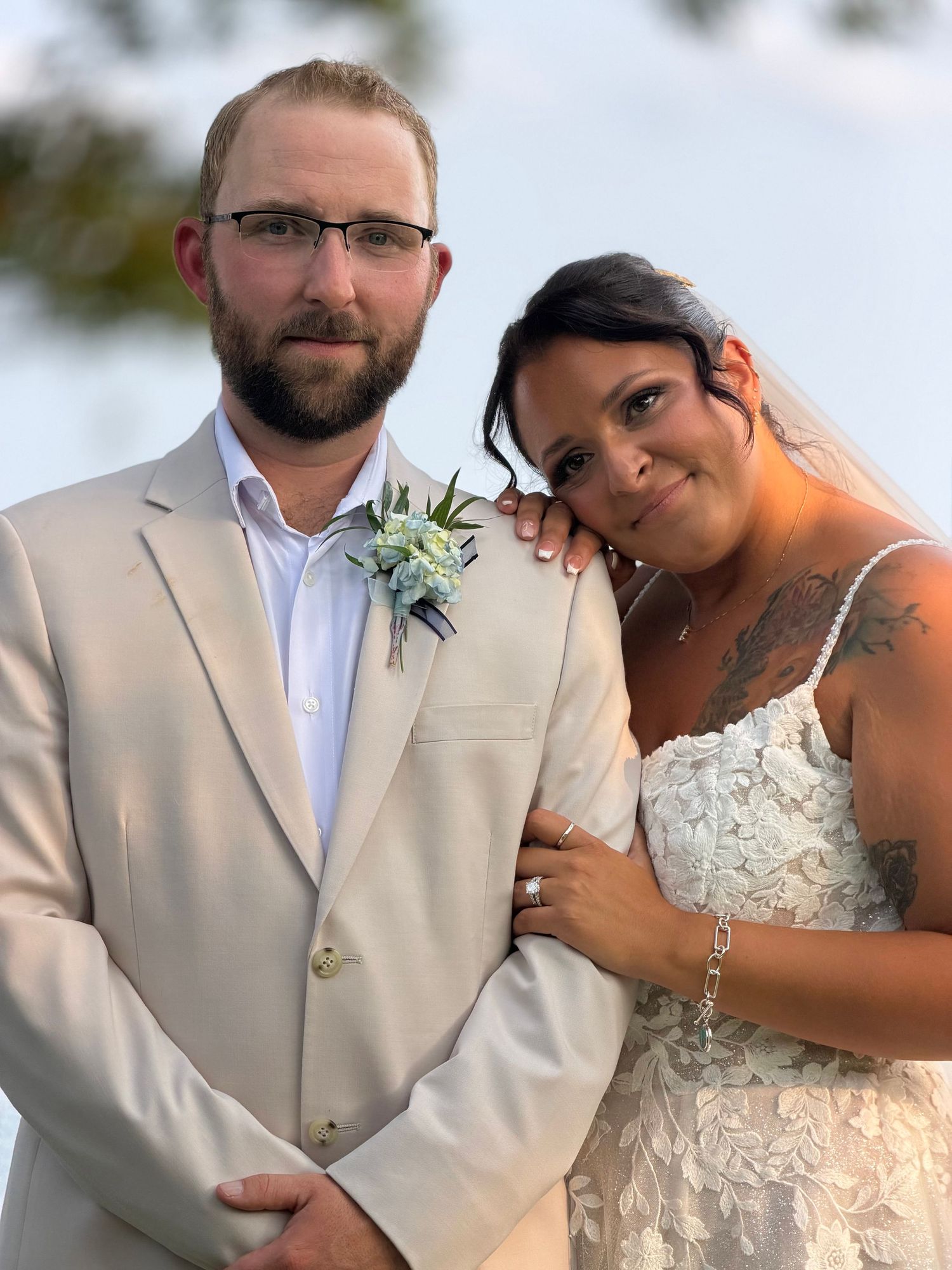 A couple in wedding attire, groom in beige suit with boutonniere, bride in lace dress with tattoos, posing outdoors.
