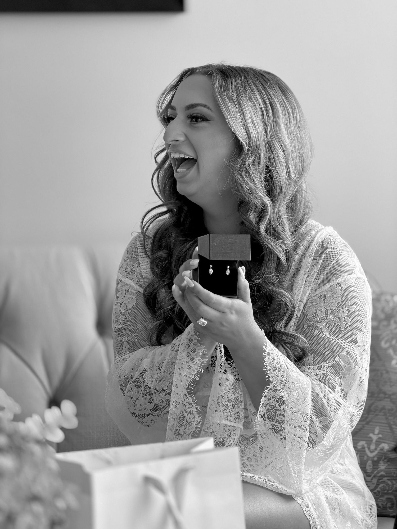 A joyful bride in lace robe holds a jewelry box with earrings, laughing during bridal preparations in black and white.