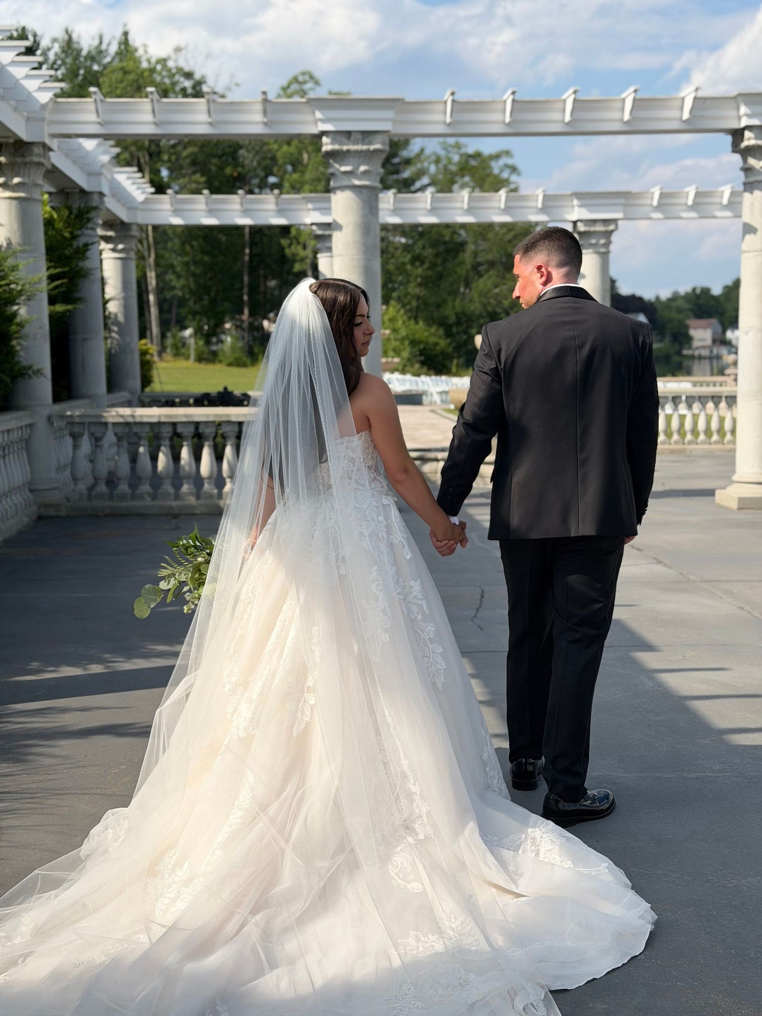 A bride in a white gown and veil holds hands with a groom in a black suit on an elegant outdoor terrace.