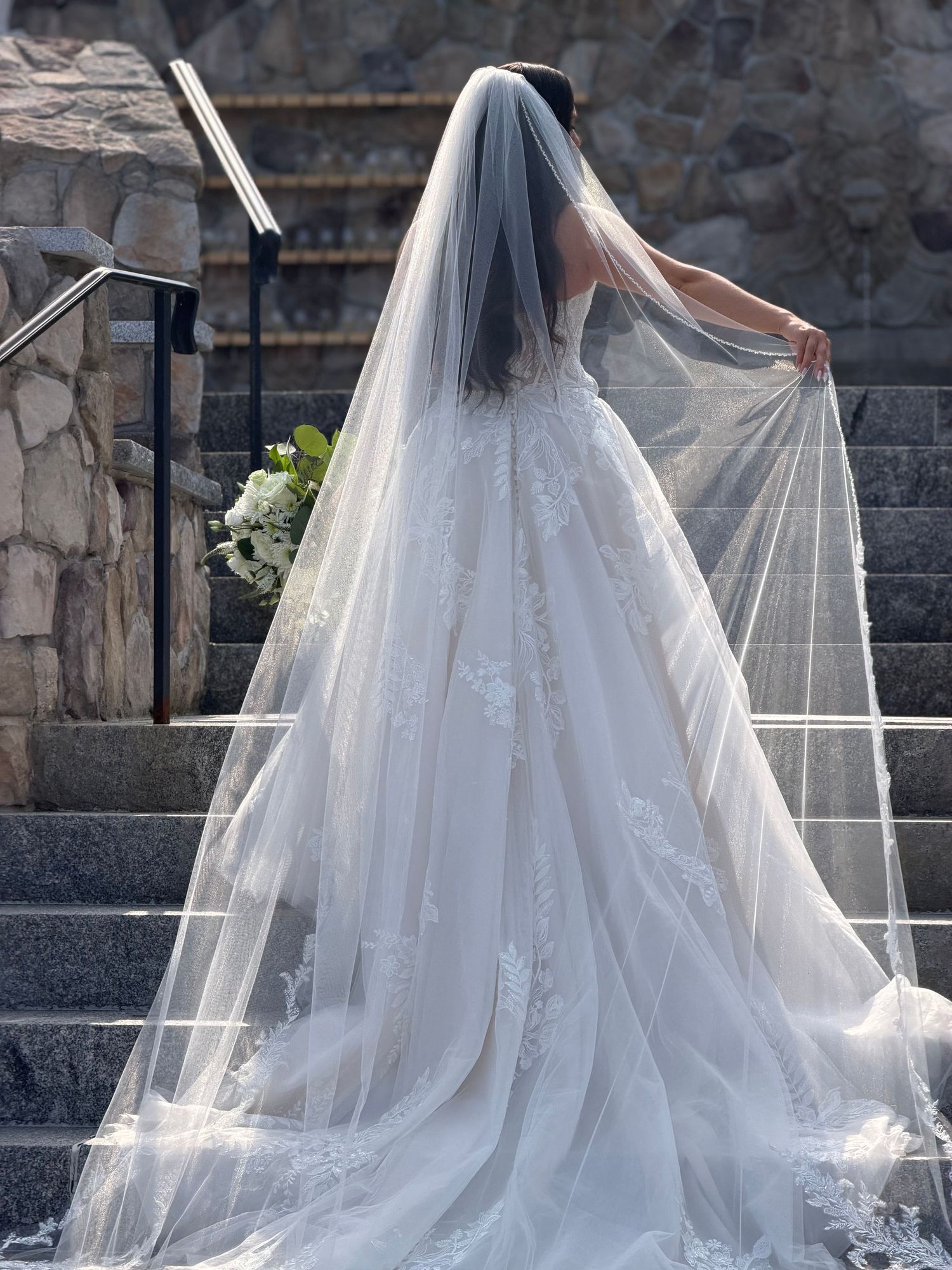 Bride in lace ball gown wedding dress with long veil ascending stone steps holding bouquet.