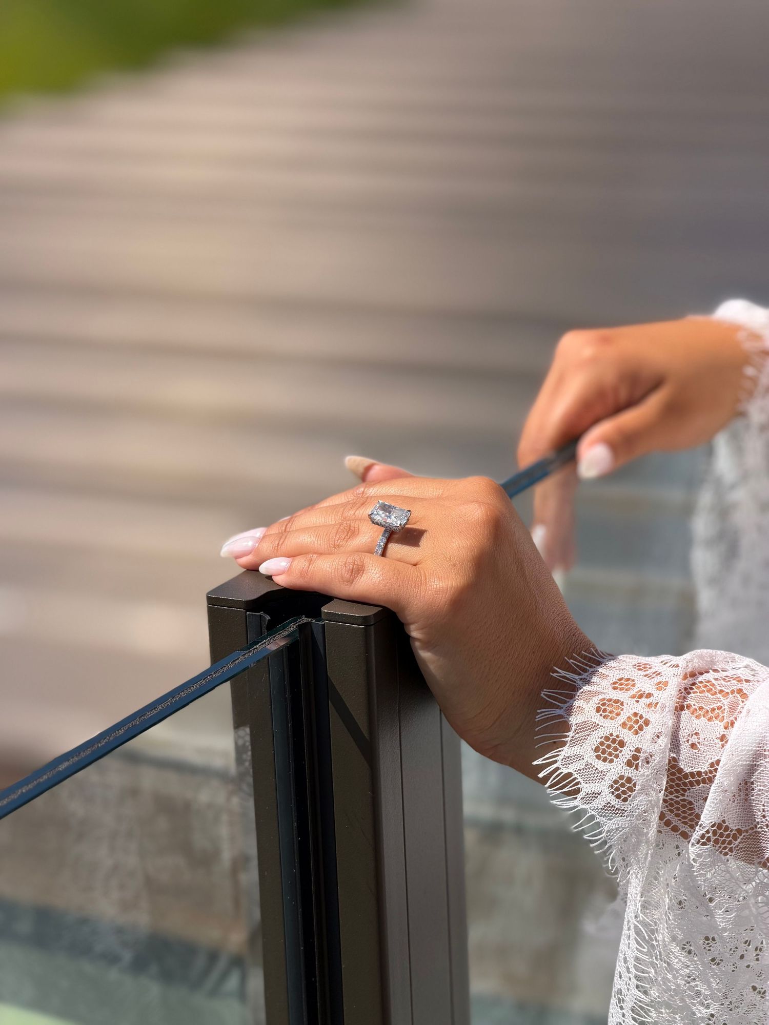 Close-up of a woman's hand with white nails and a diamond ring, holding a pen on a glass railing.