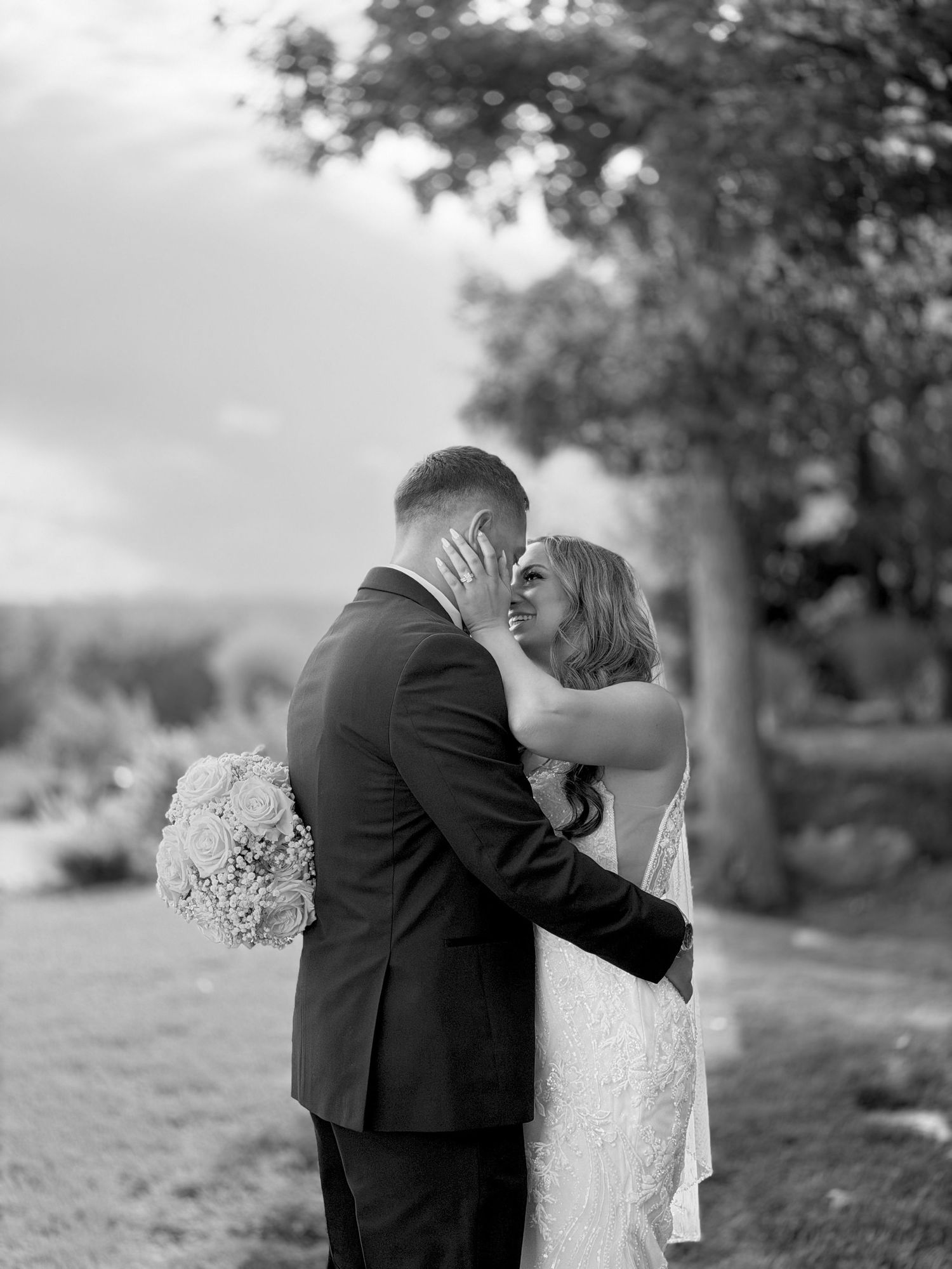 Black and white photo of a bride and groom embracing and smiling at each other outdoors on their wedding day.