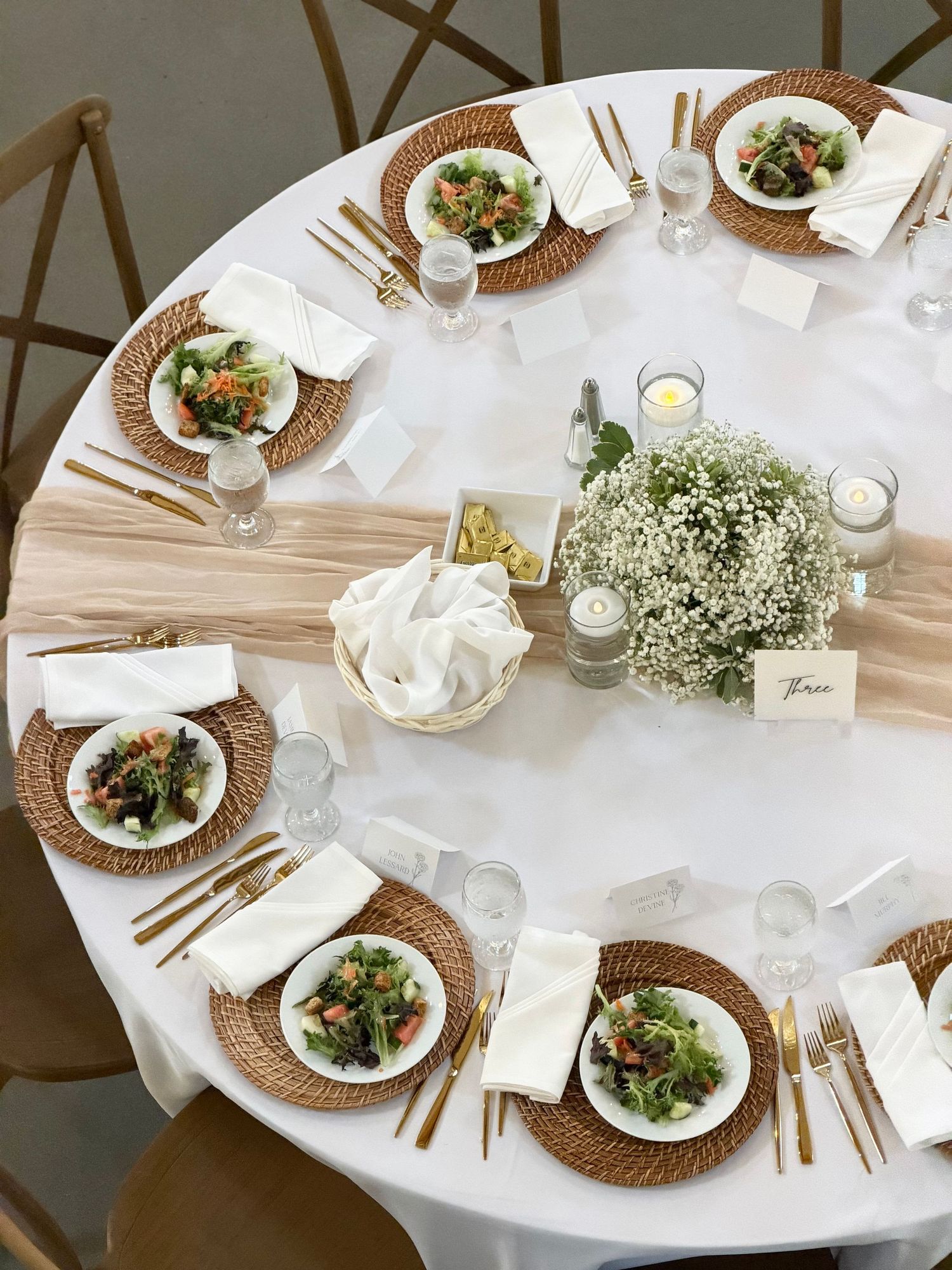 Elegant round wedding table with wicker chargers, gold cutlery, white napkins, baby's breath centerpiece, and fresh salad plates.