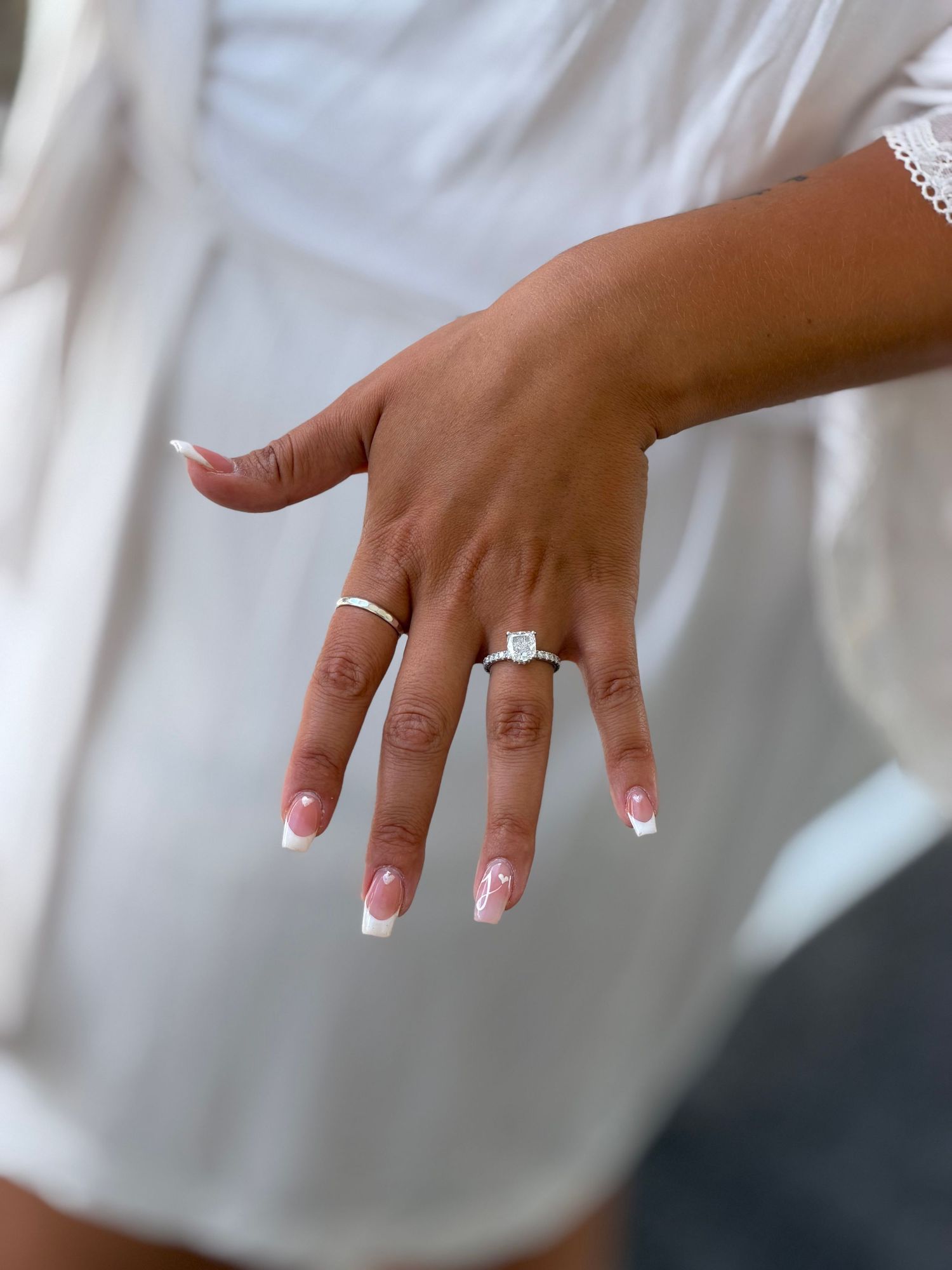 Close-up of a hand with French tip nails wearing a solitaire diamond engagement ring and a thin gold band.
