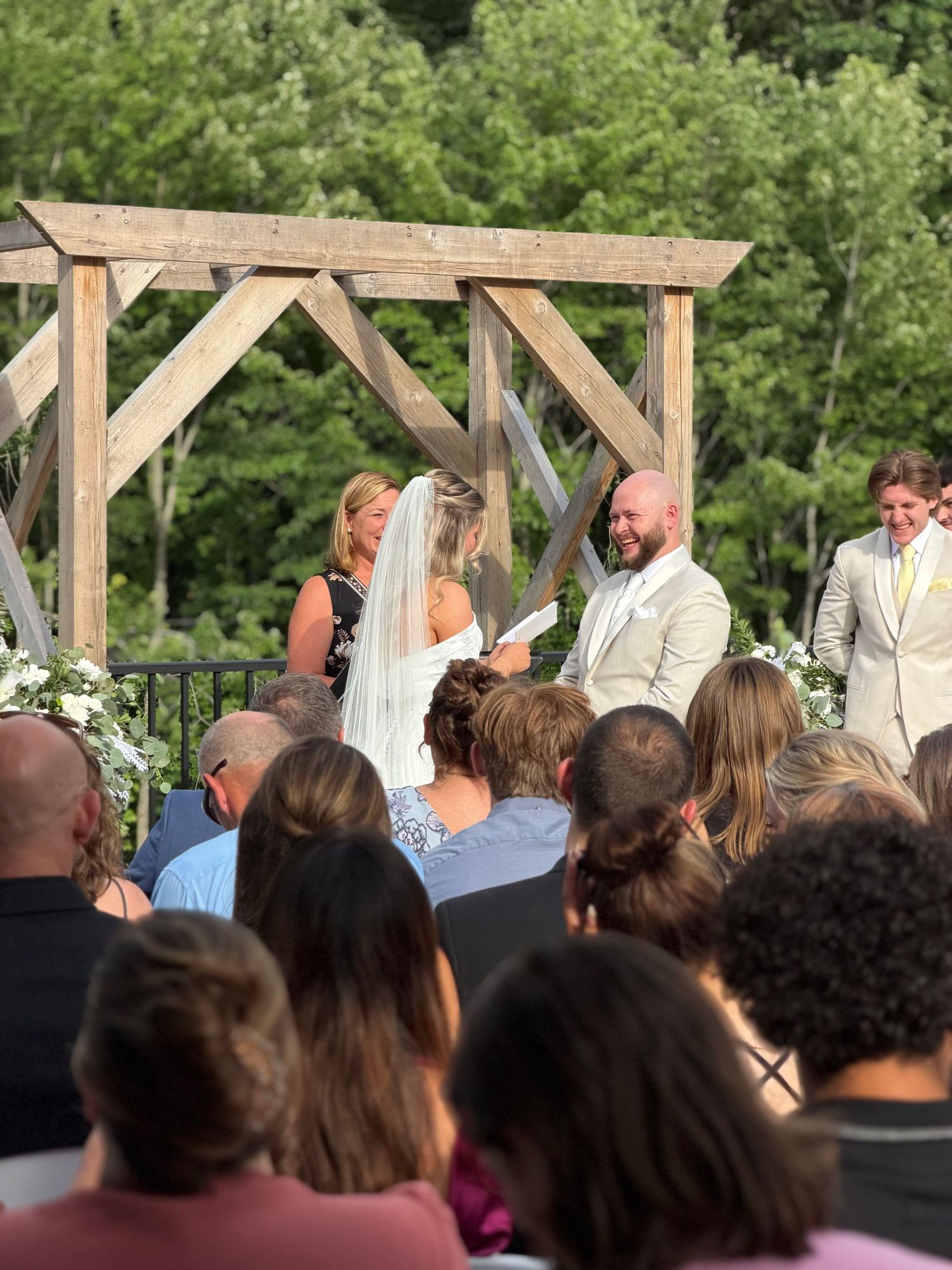 Outdoor wedding ceremony with bride and groom at rustic wooden arch, officiant reading vows, guests seated in foreground.