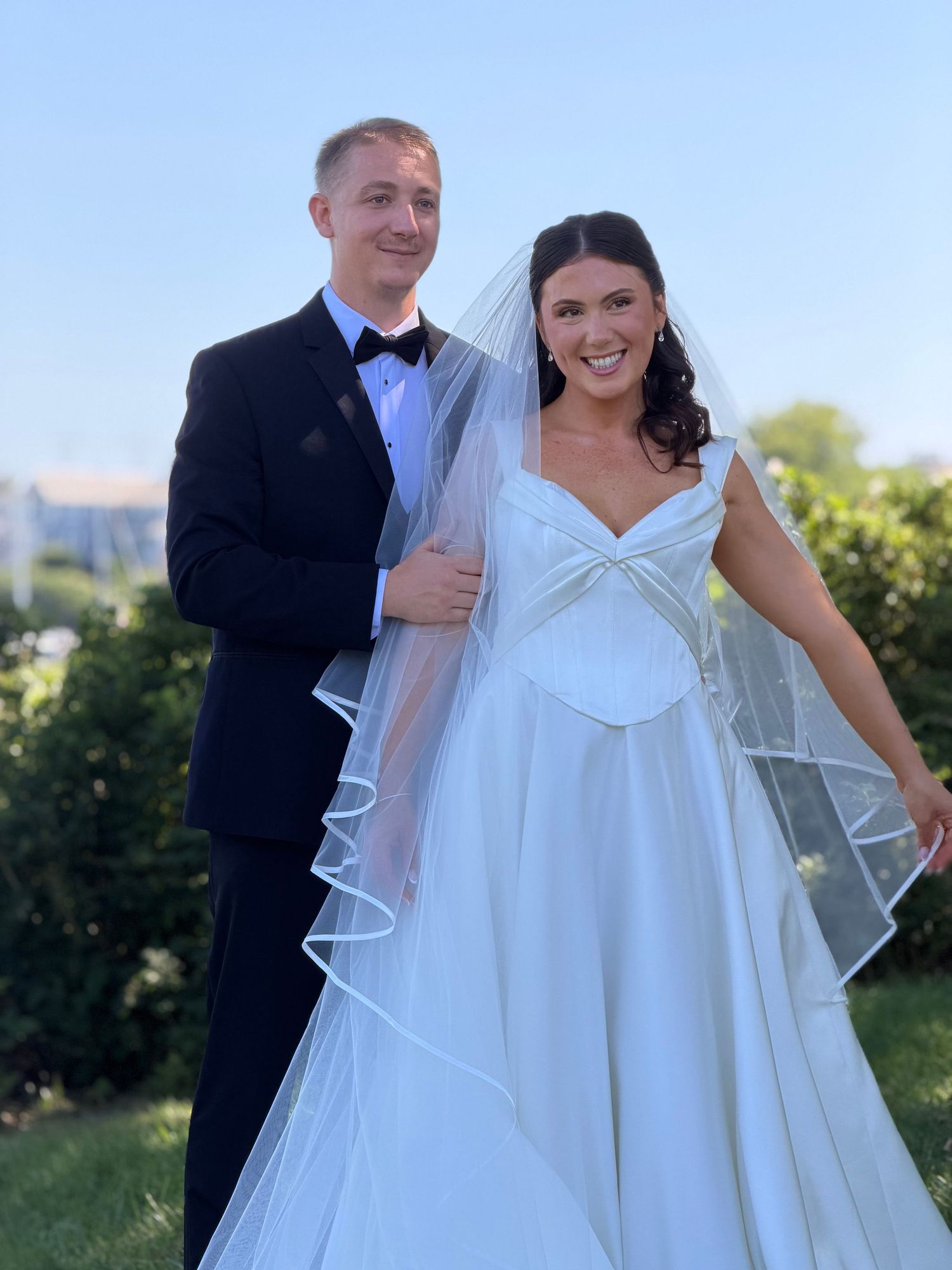 A smiling bride in a white gown and veil poses with a groom in a black tuxedo outdoors on a sunny day.
