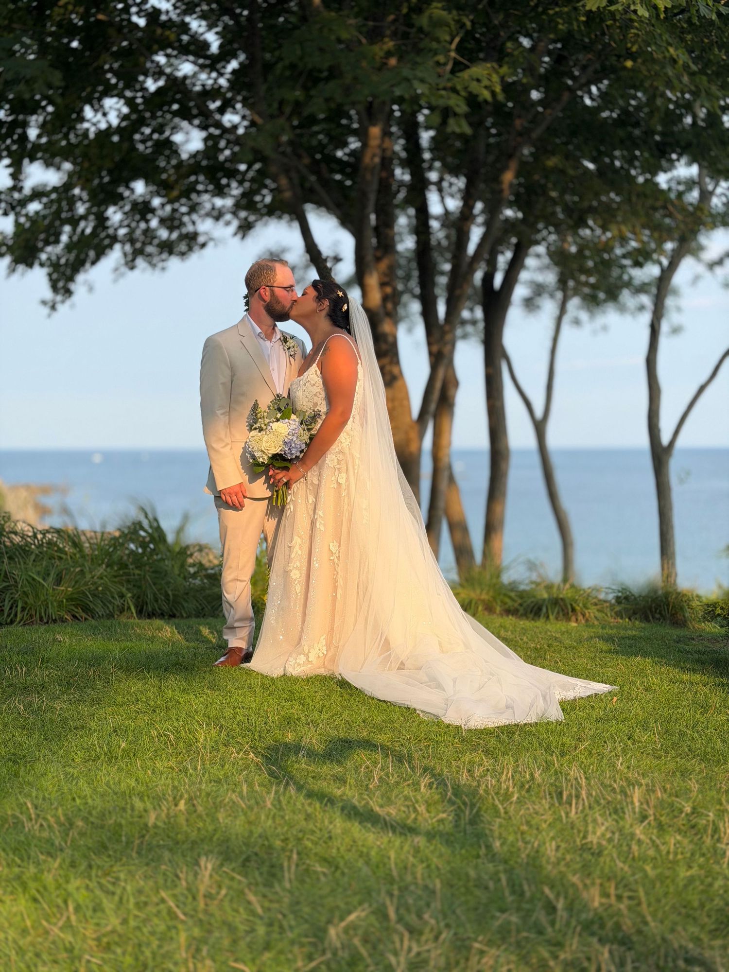 A bride and groom share a romantic kiss on a green lawn with trees and ocean views during their outdoor wedding ceremony.