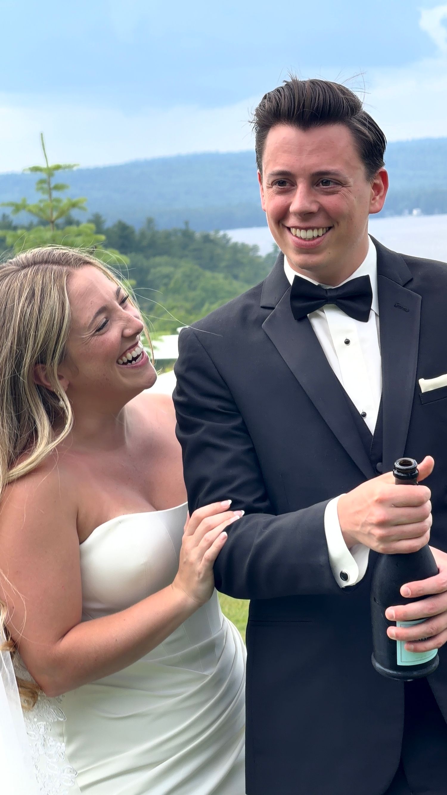 Happy newlyweds celebrate with champagne, bride in white gown and groom in black tuxedo, scenic lake backdrop.