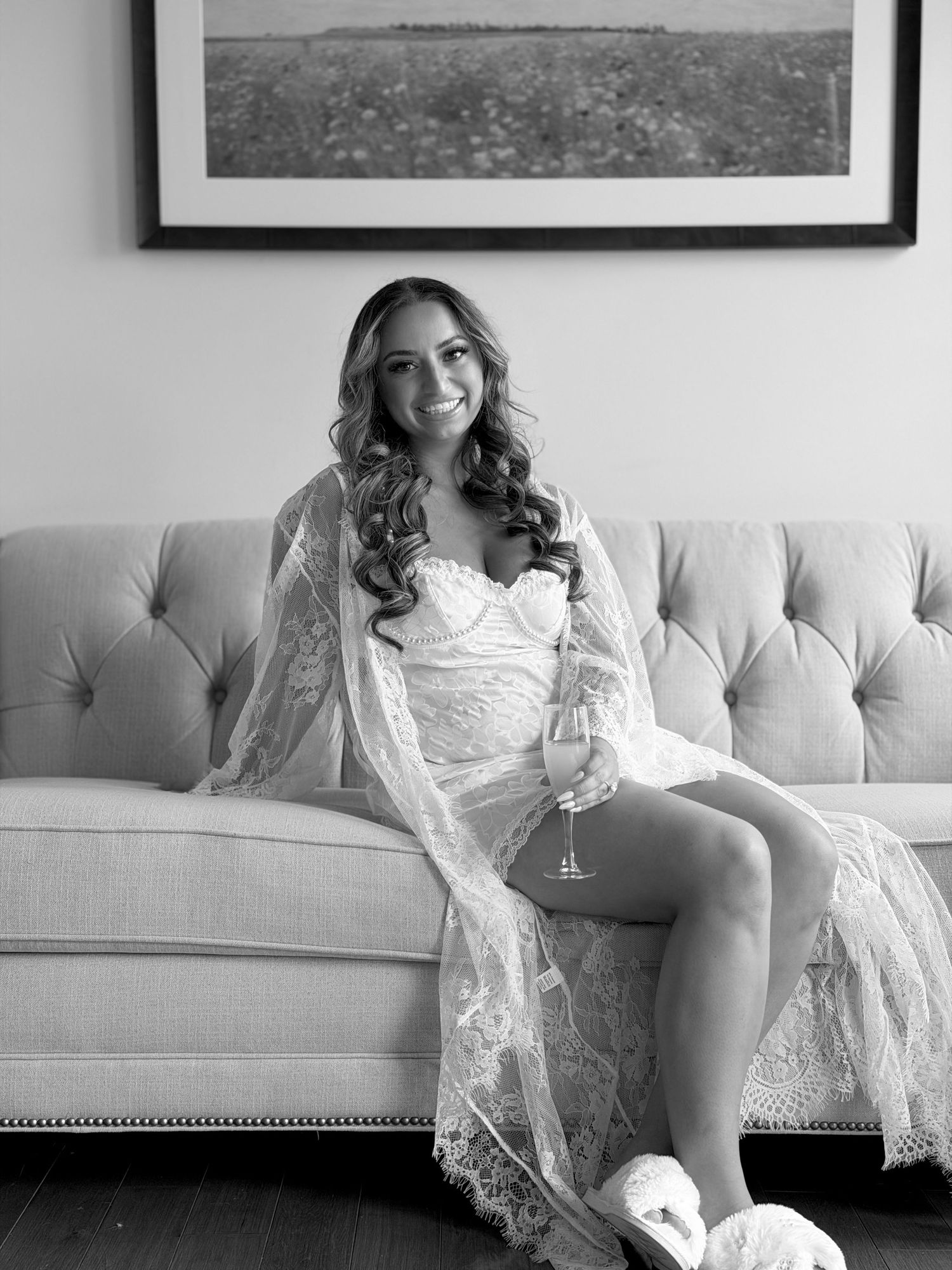 Black and white photo of a smiling bride in lace robe holding champagne, seated on a tufted sofa wearing fluffy slippers.
