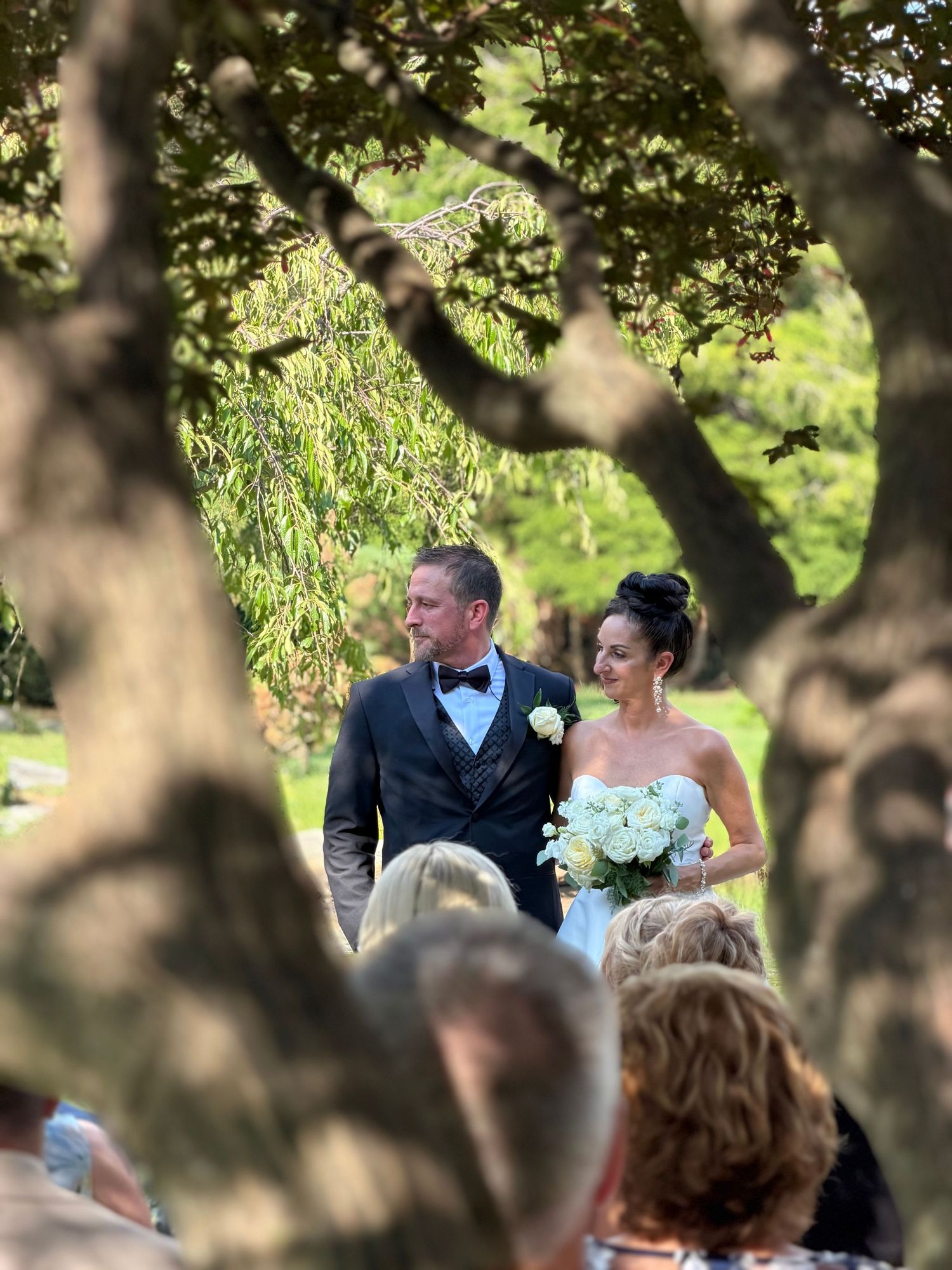A bride and groom stand together outdoors under lush green trees during an elegant garden wedding ceremony.