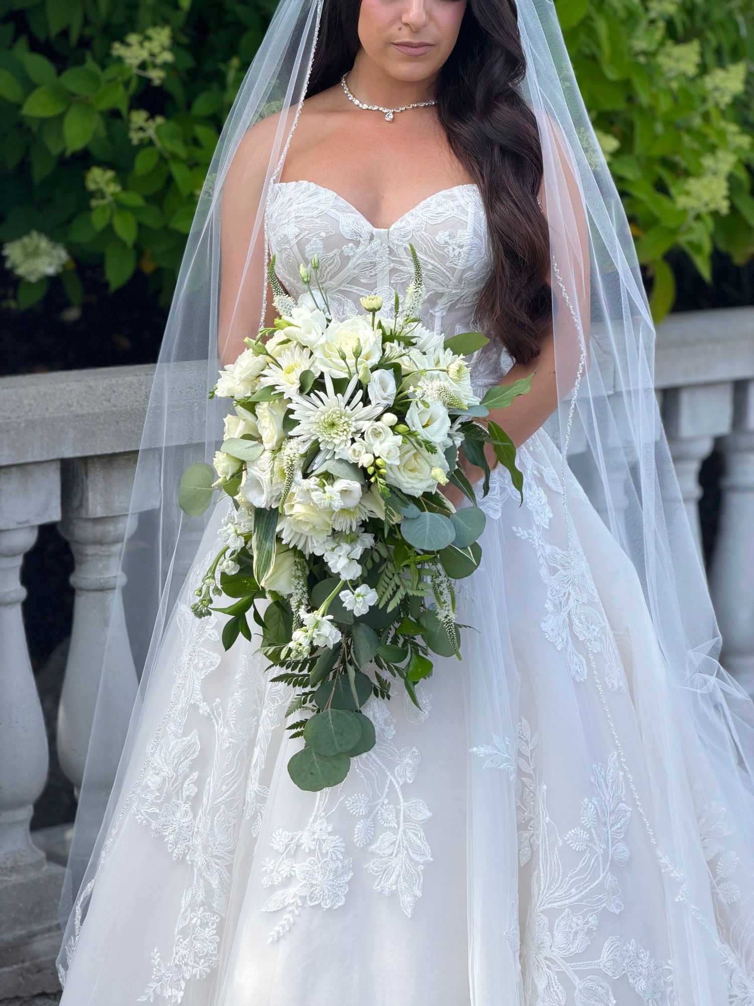 Bride in strapless lace wedding gown with veil holding cascading white floral bouquet with greenery outdoors.