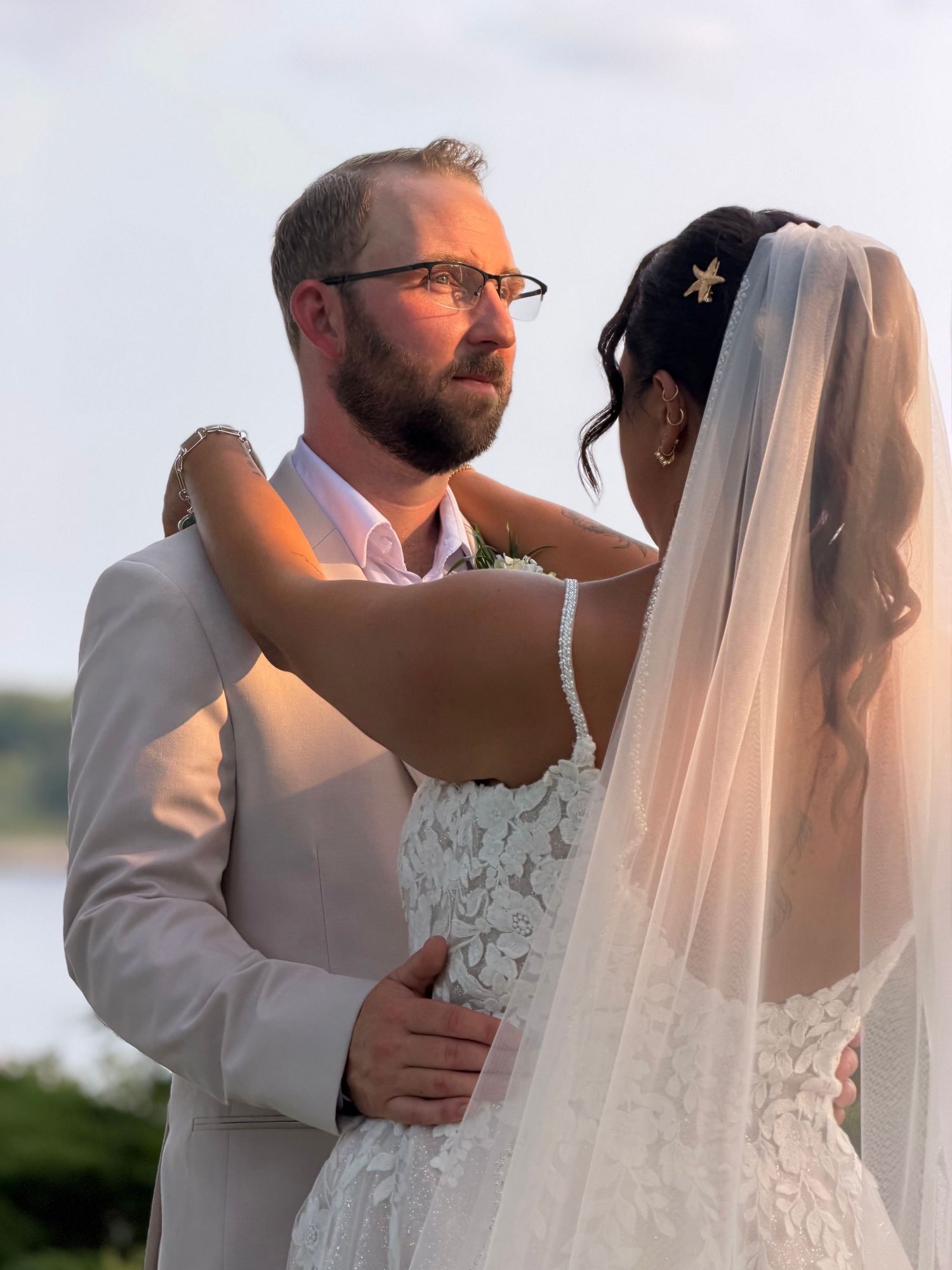 A bride in a white lace gown and veil embraces a bearded groom in a tan suit during an outdoor wedding ceremony.