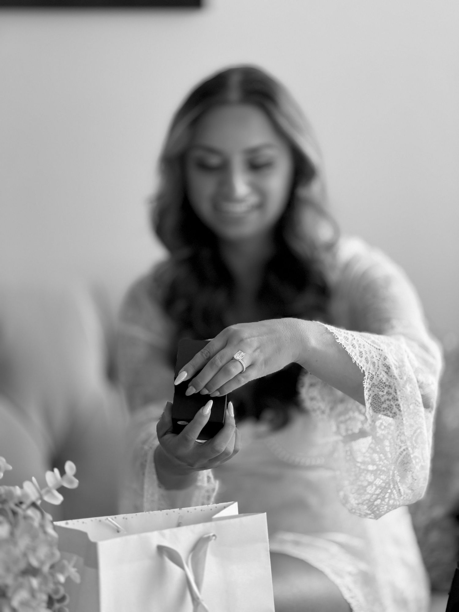 Black and white photo of a smiling bride in lace robe examining her engagement ring with a gift bag in foreground.