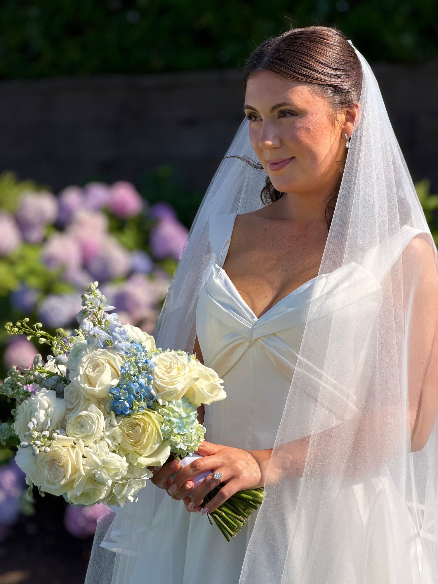 Bride in white gown and veil holding blue and white rose bouquet, smiling outdoors near blooming hydrangeas.