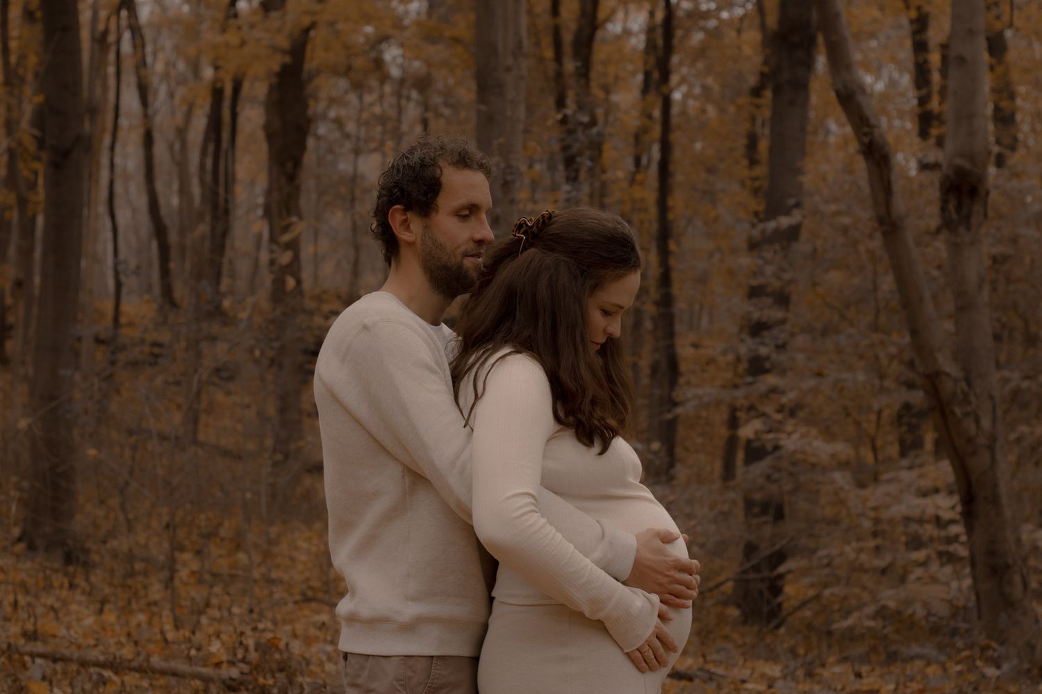 Séance photo couple à Montréal, étreinte en pulls crème dans une forêt automnale aux feuilles dorées et brunes.