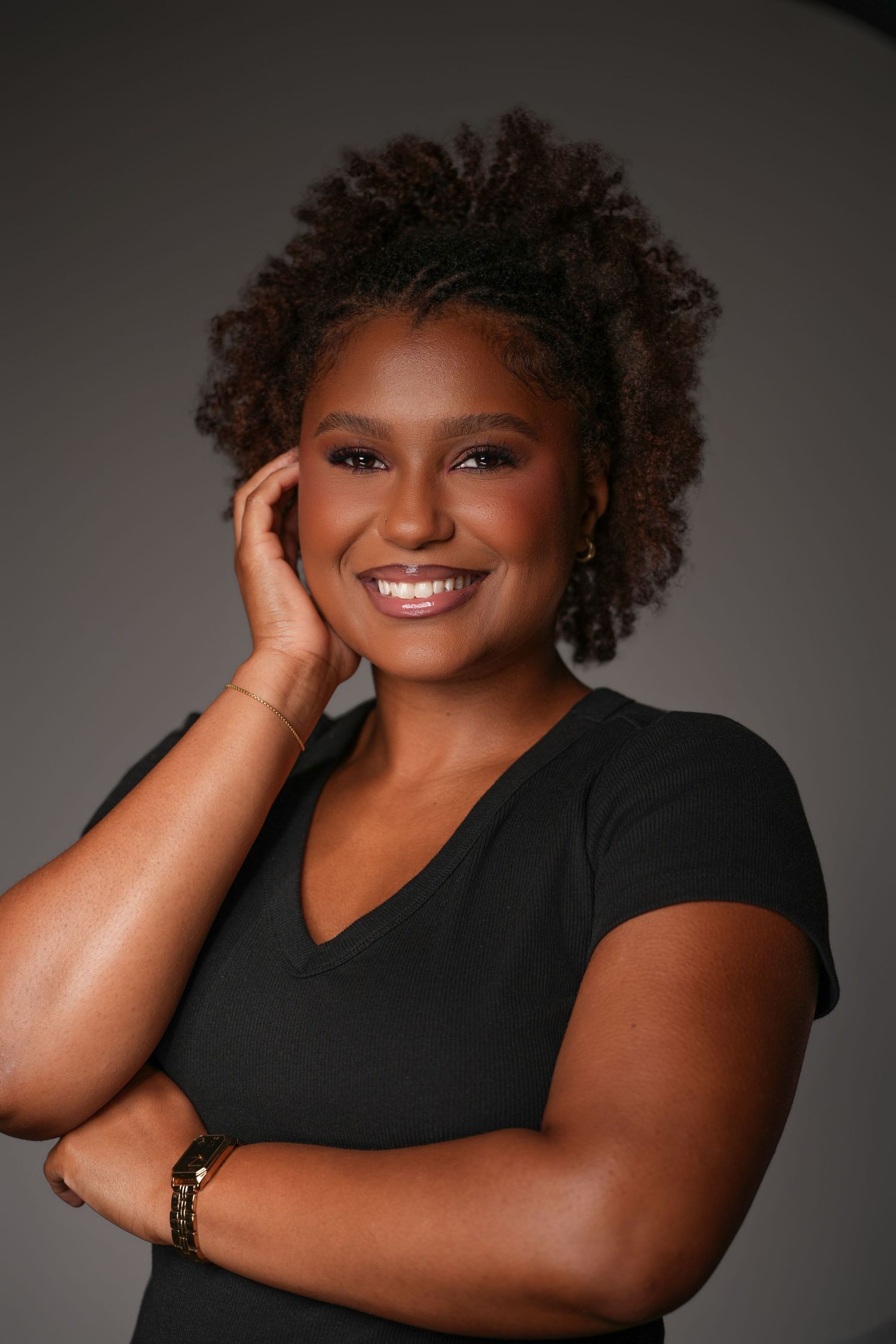 Smiling woman with short natural curly hair poses with one hand at her face and arms crossed, wearing a black v-neck top.