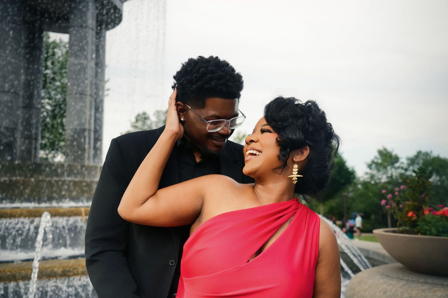 Smiling couple embracing in front of a fountain, the woman in a red satin dress and gold earrings, the man in black with glasses.