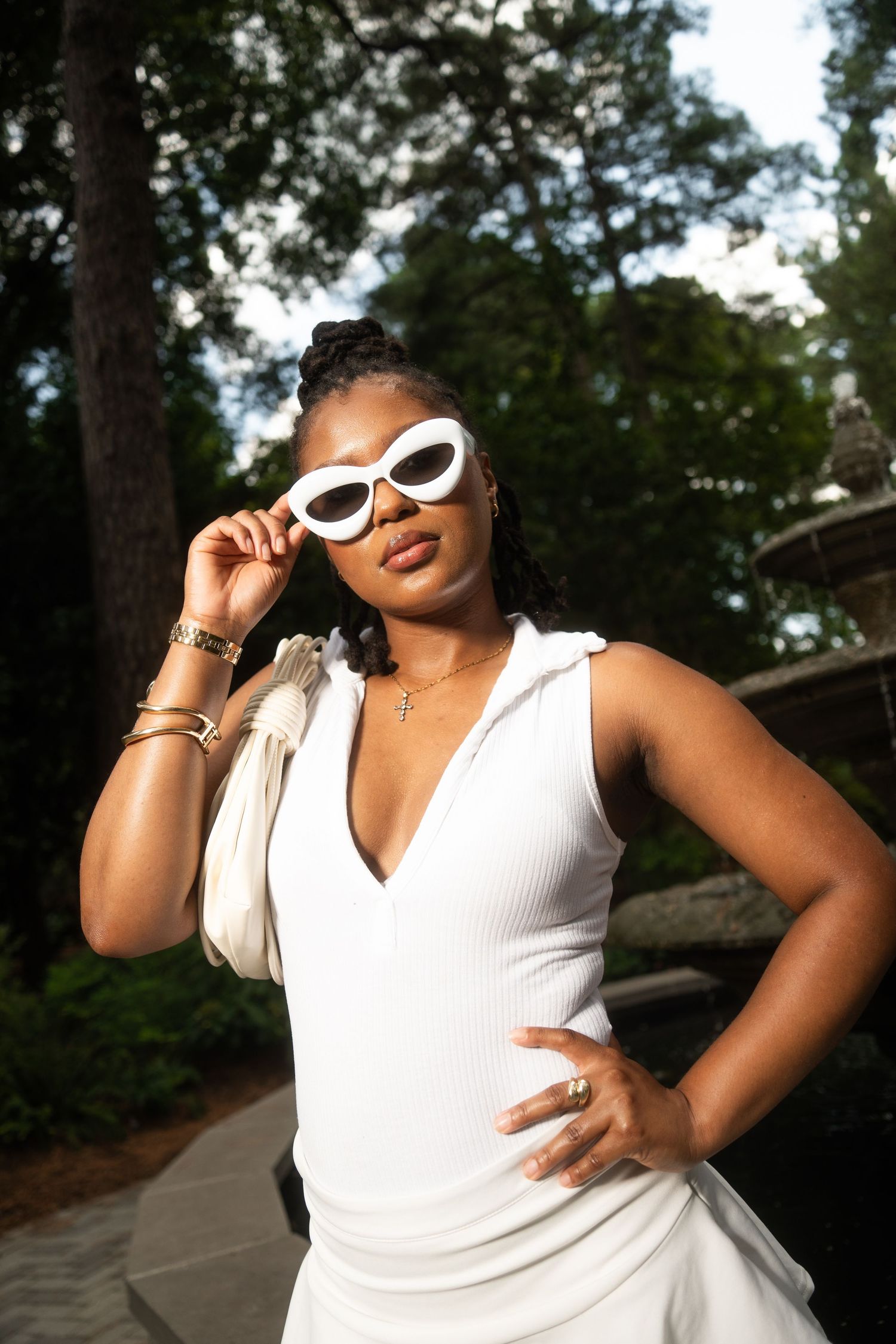 Woman in a white sleeveless top and white sunglasses poses outdoors by a stone fountain, with gold bracelets and a cross necklace.