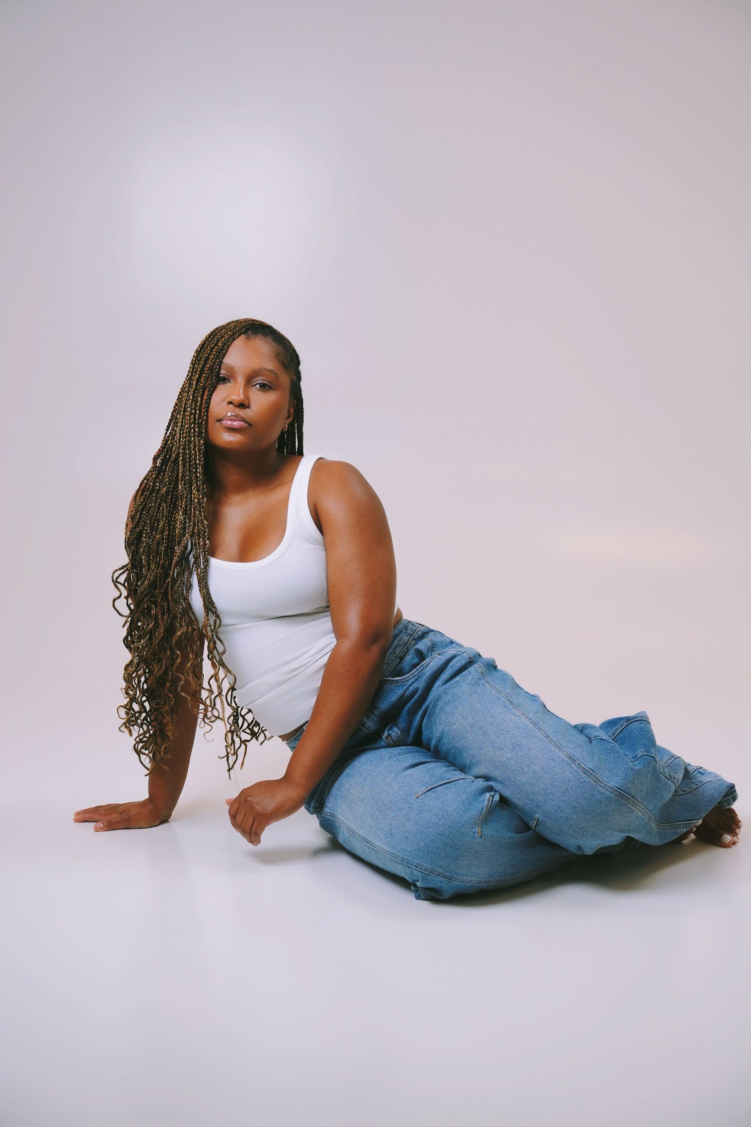Woman with long braids sitting on a white studio floor in jeans and white tank top.
