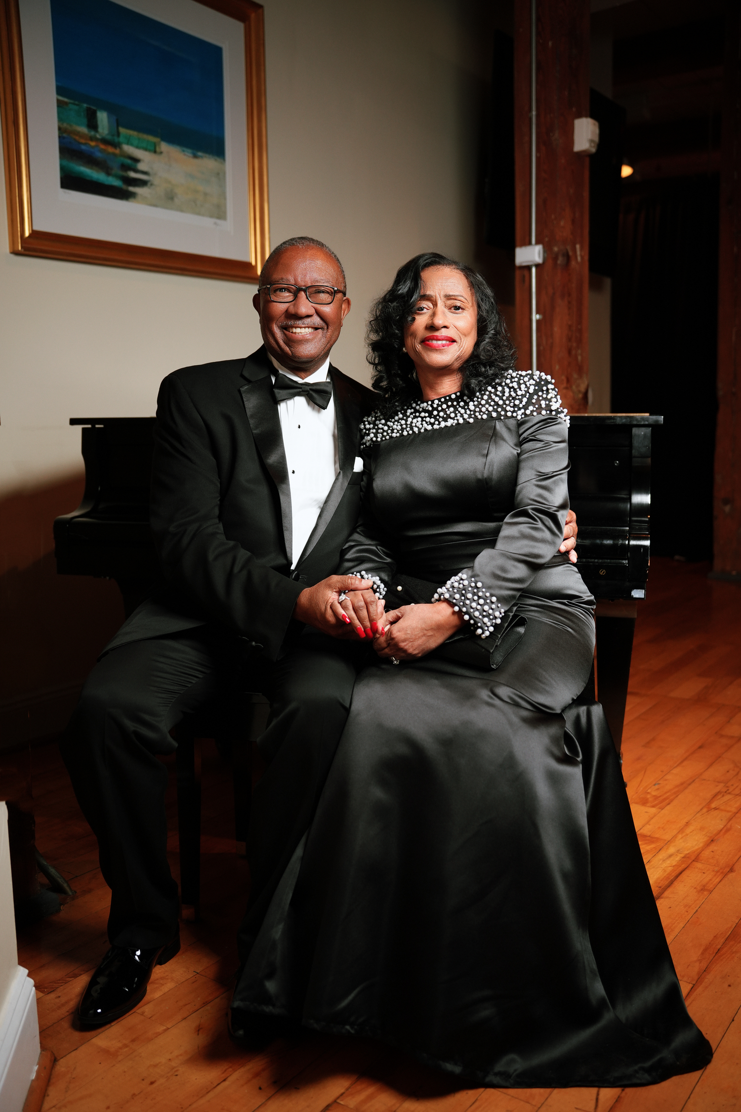 Smiling couple in formal evening wear sit together on a piano bench, he in a black tuxedo and bow tie, she in a black satin gown.