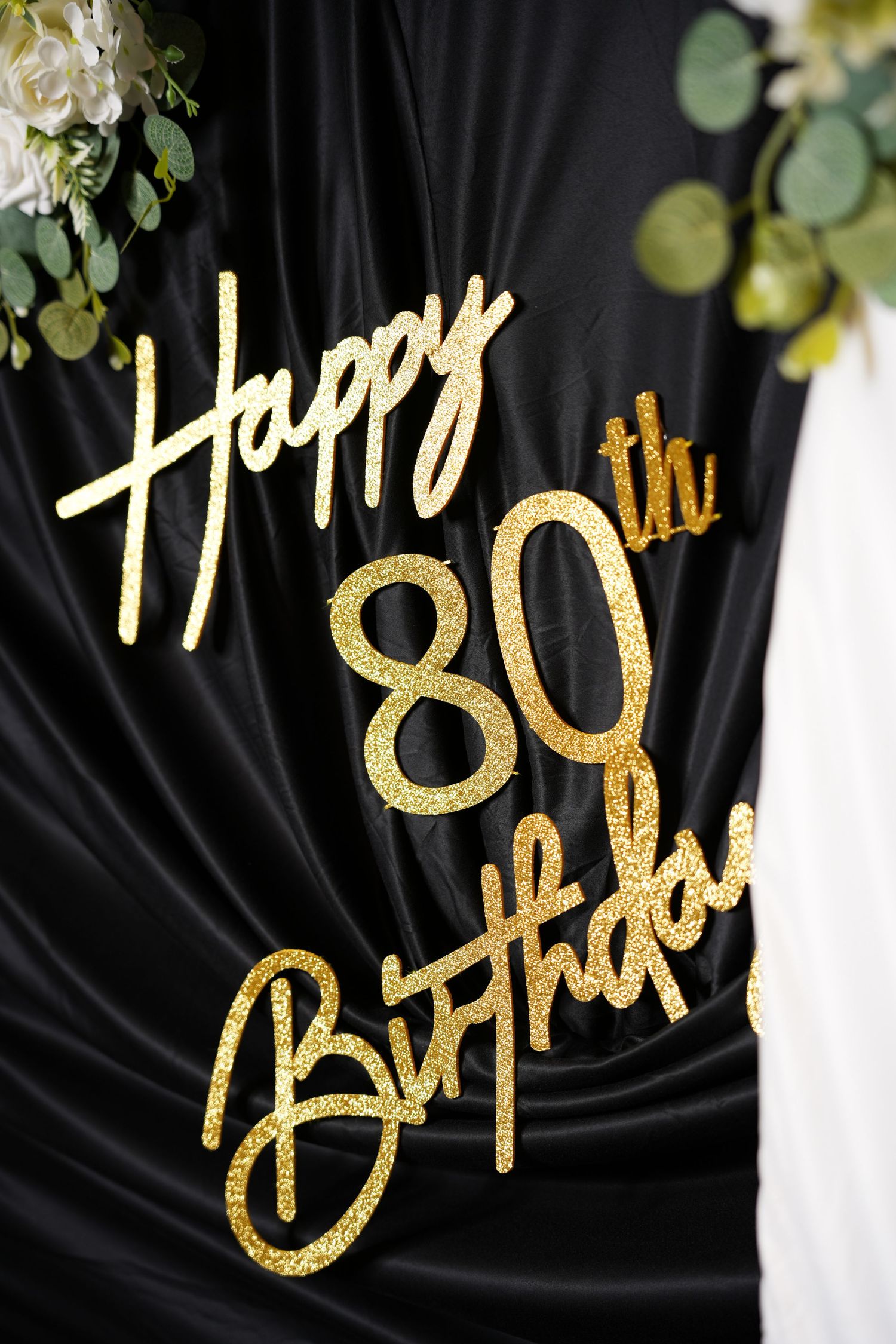 Gold glitter Happy 80th Birthday sign on black draped fabric backdrop with white flowers and green eucalyptus decor.