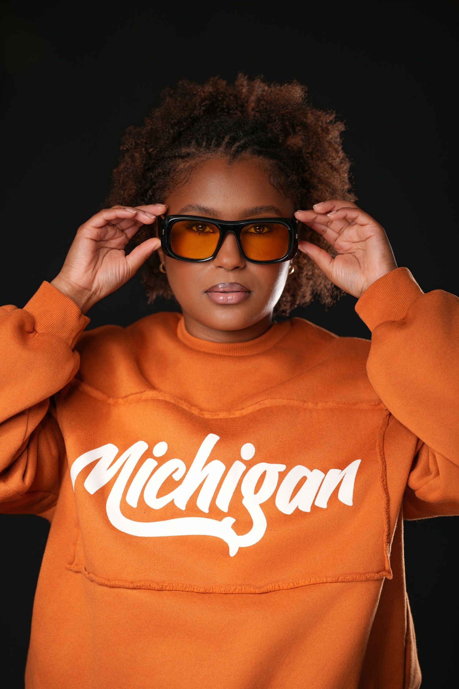 Woman in orange-tinted sunglasses and a rust orange Michigan script sweatshirt poses against a black background.