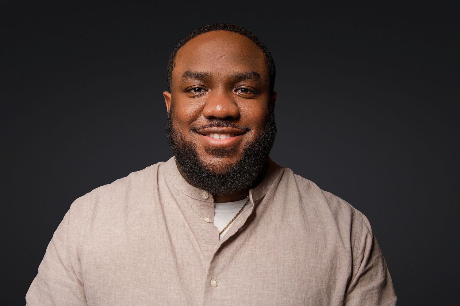 Smiling man with a full beard wearing a beige button-collar shirt, posed against a dark gray studio background.