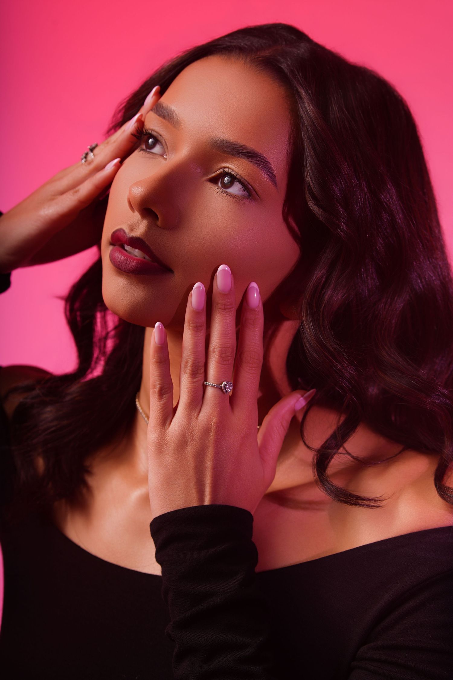 Elegant woman with wavy dark hair poses with hands near face, showcasing a diamond ring against a pink backdrop.