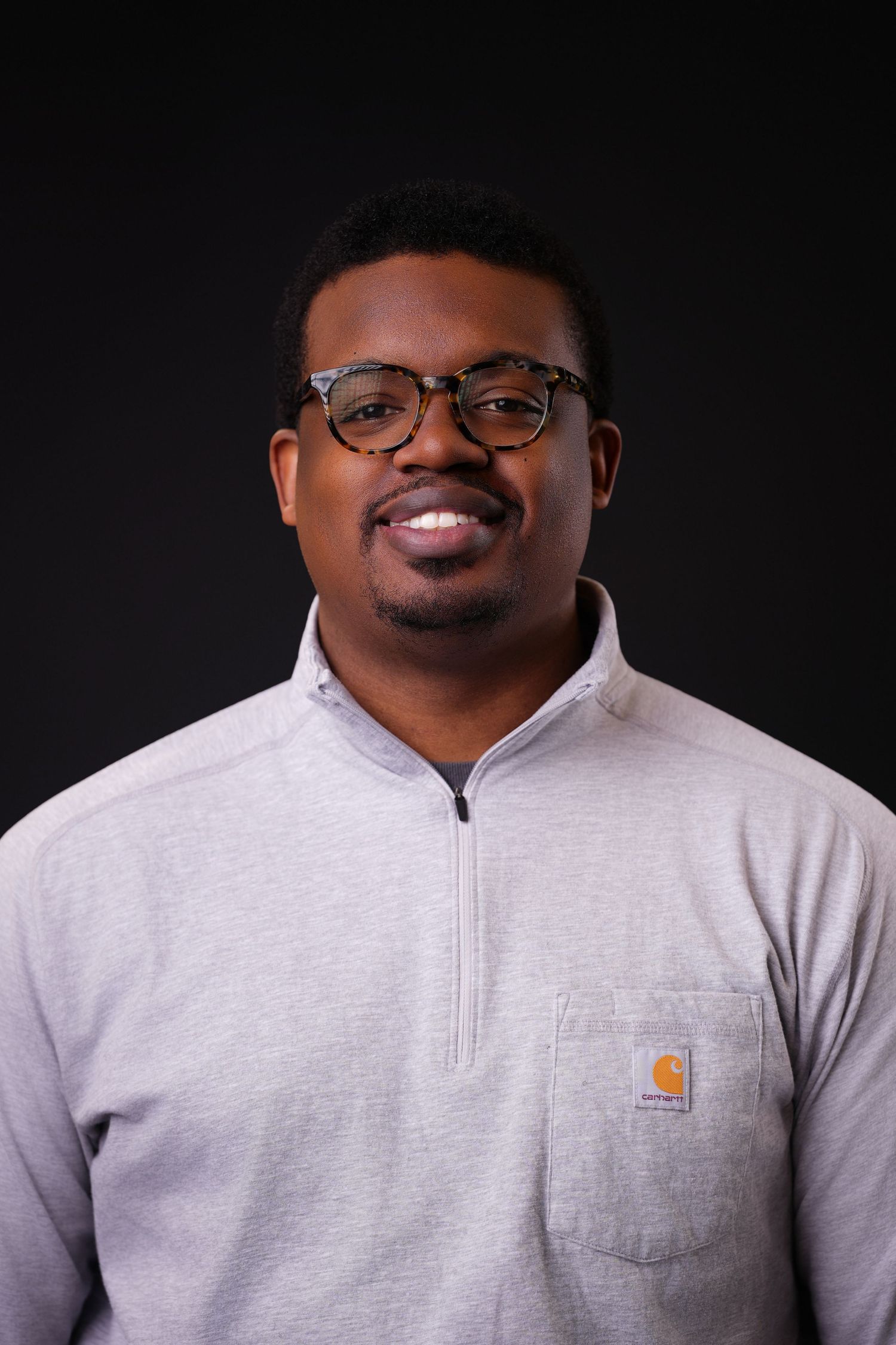 Smiling man with short dark hair, tortoiseshell glasses and a goatee wears a gray quarter-zip Carhartt pullover against a dark background.