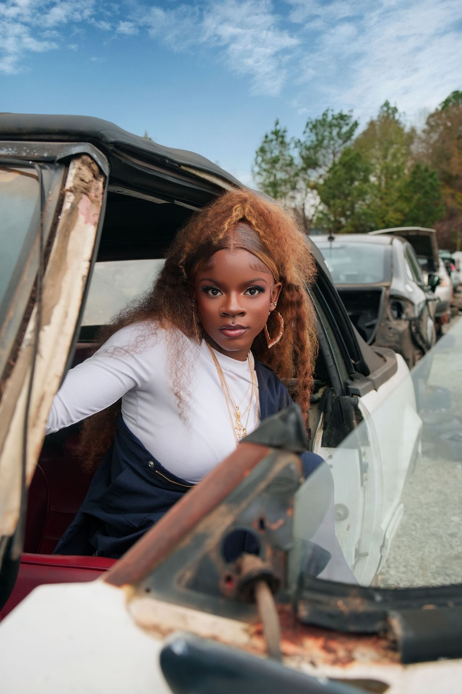 A young Black woman with curly auburn hair poses beside a rusty vintage car in a junkyard.