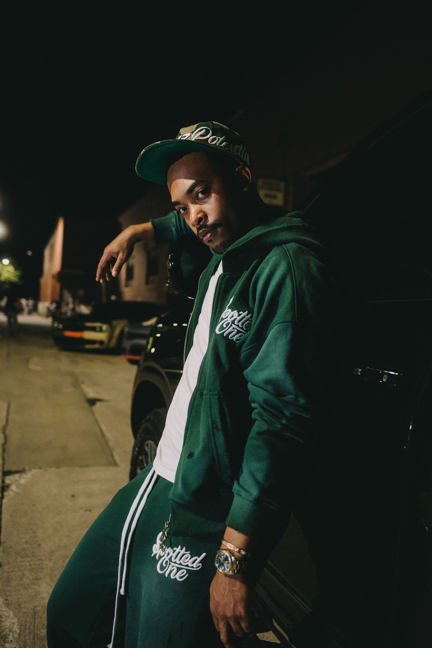 Man in a green hoodie, matching sweatpants and a snapback cap leans against a dark car on a night street, wearing a watch and bracelet.