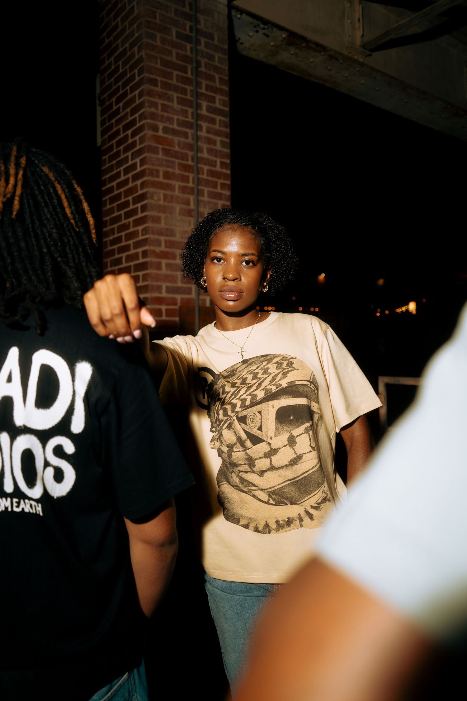 A young Black woman in a graphic tee stands among a crowd near a brick wall at night.