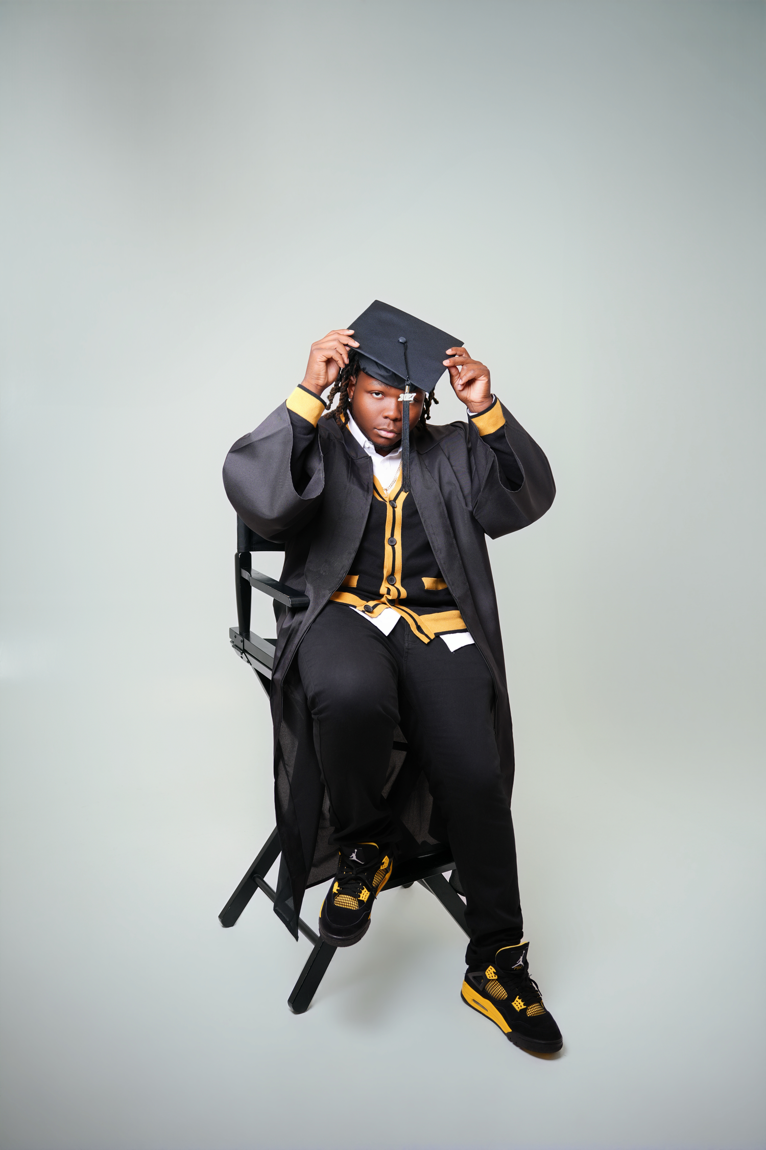 Graduate in a black gown and mortarboard sits on a director's chair, adjusting the cap, wearing black and gold sneakers.
