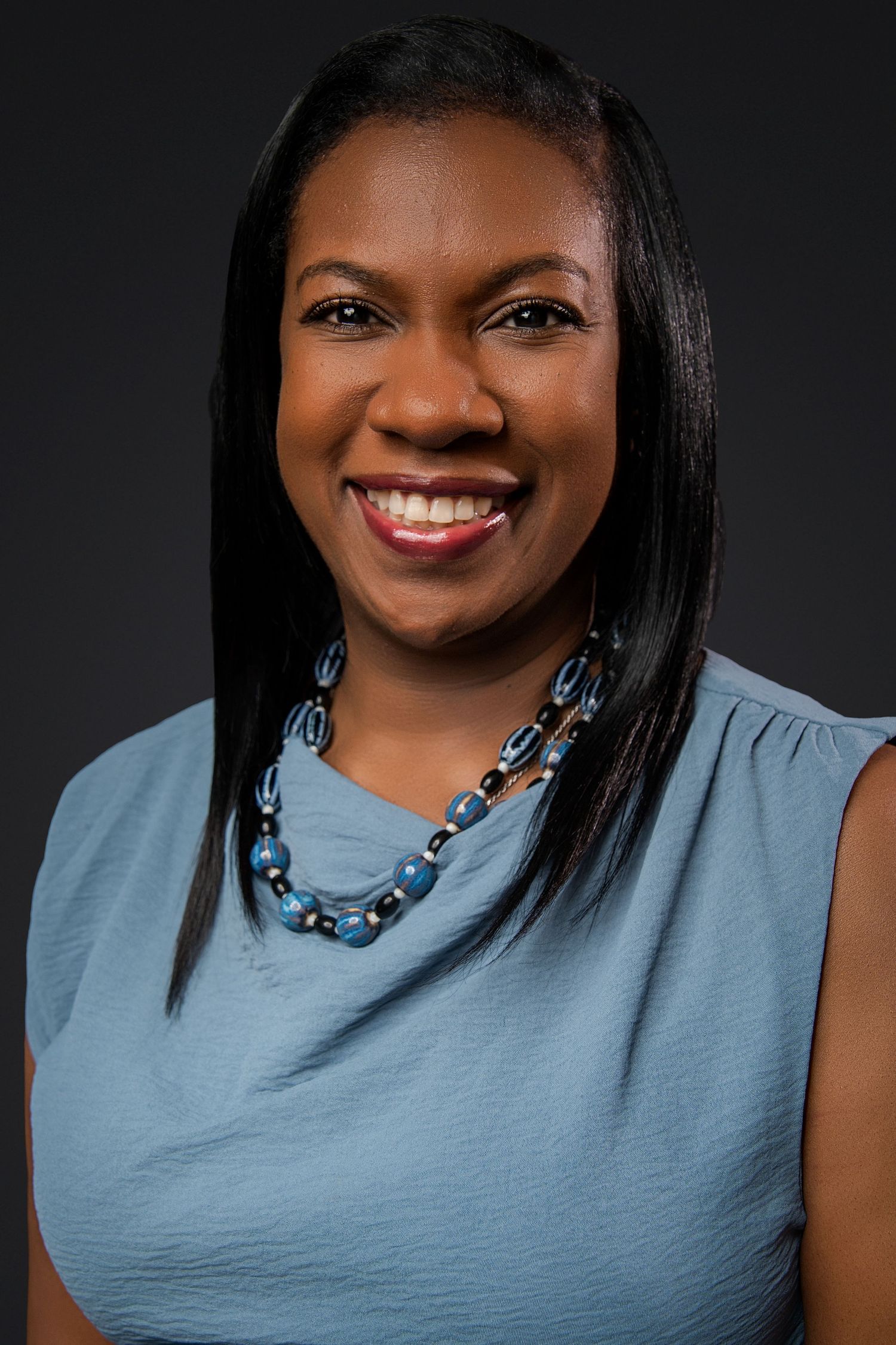 Smiling woman with long straight black hair wearing a blue top and a blue beaded necklace poses against a dark background.