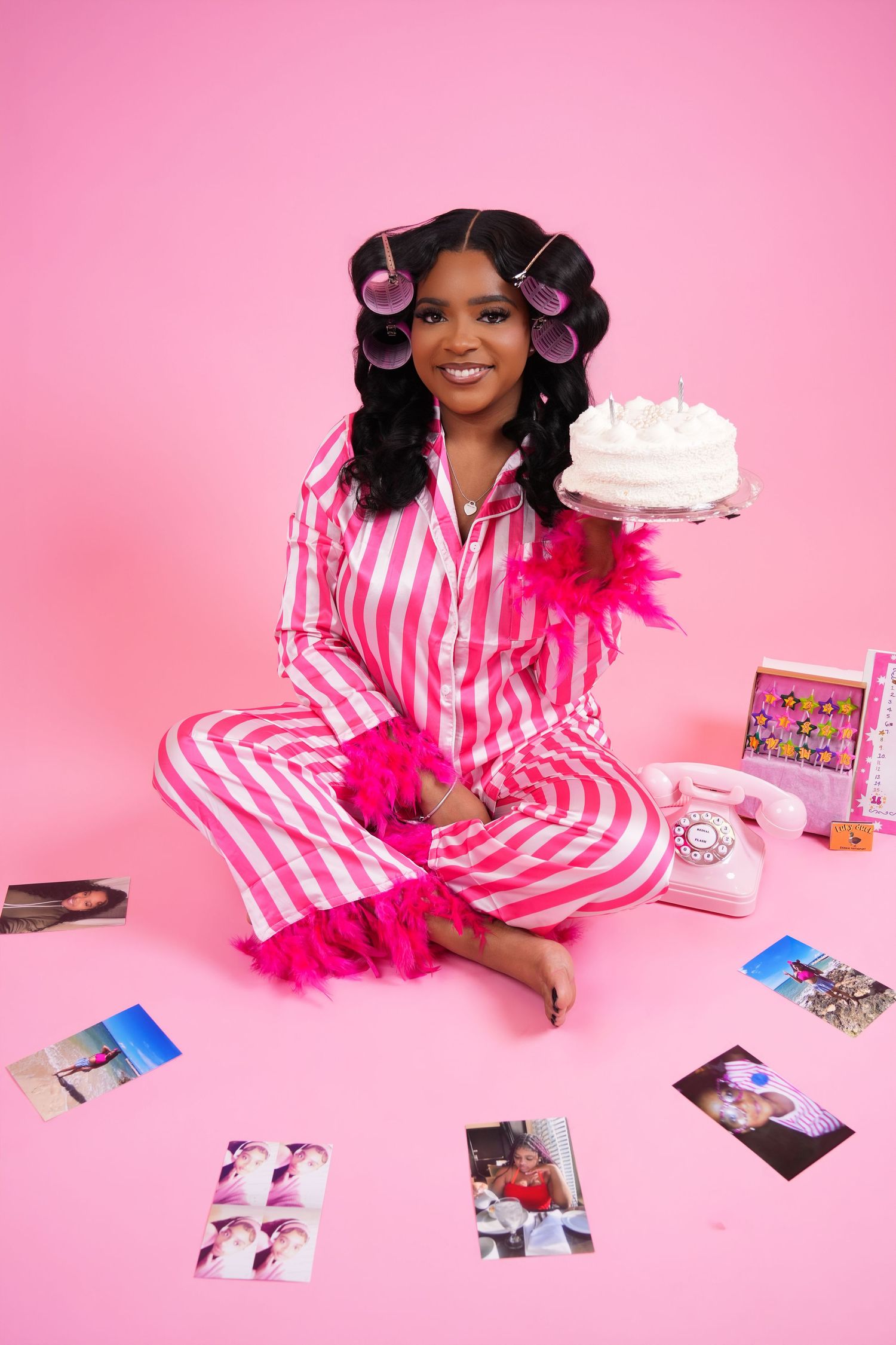 Woman in pink striped pajamas sits on pink floor holding a birthday cake surrounded by scattered photos.