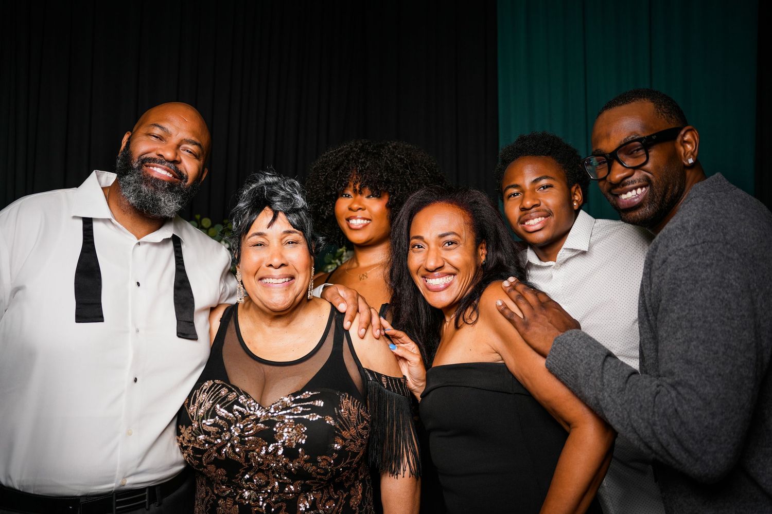 Six smiling people in elegant evening attire pose close together with arms around each other against a dark backdrop at a festive event.